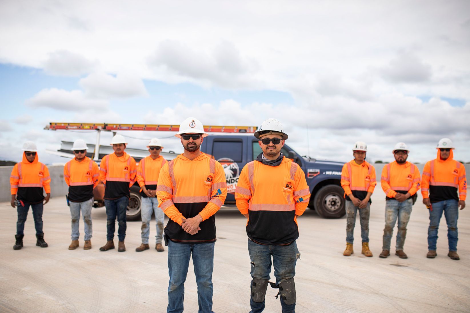 Workers in orange safety shirts stand in front of heavy equipment under a cloudy sky.