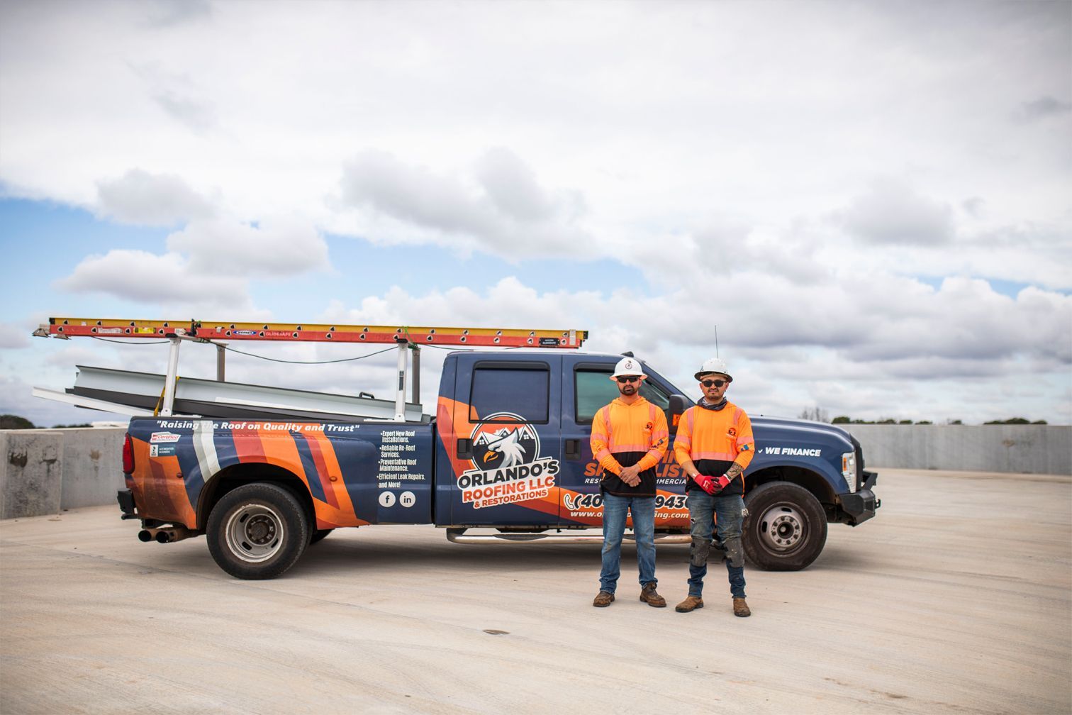 Two utility workers in orange vests stand beside a blue service truck with a ladder on a cloudy day.
