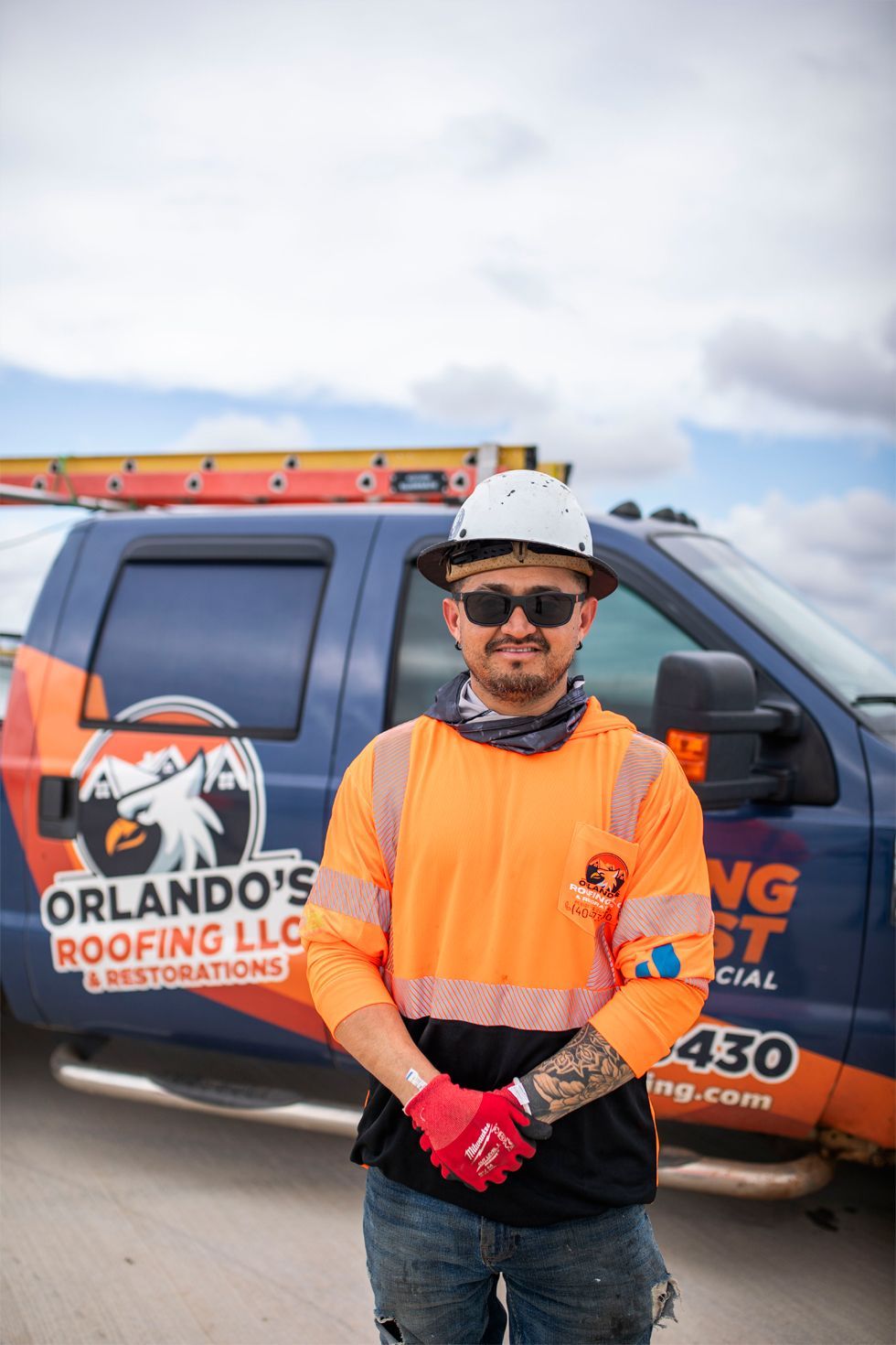 Roofing worker in orange safety vest stands beside Orlando’s Roofing LLC truck under a cloudy sky