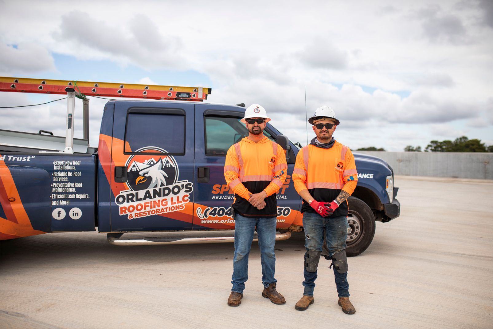 Two workers in orange safety shirts stand beside an Orlando truck under a cloudy sky.