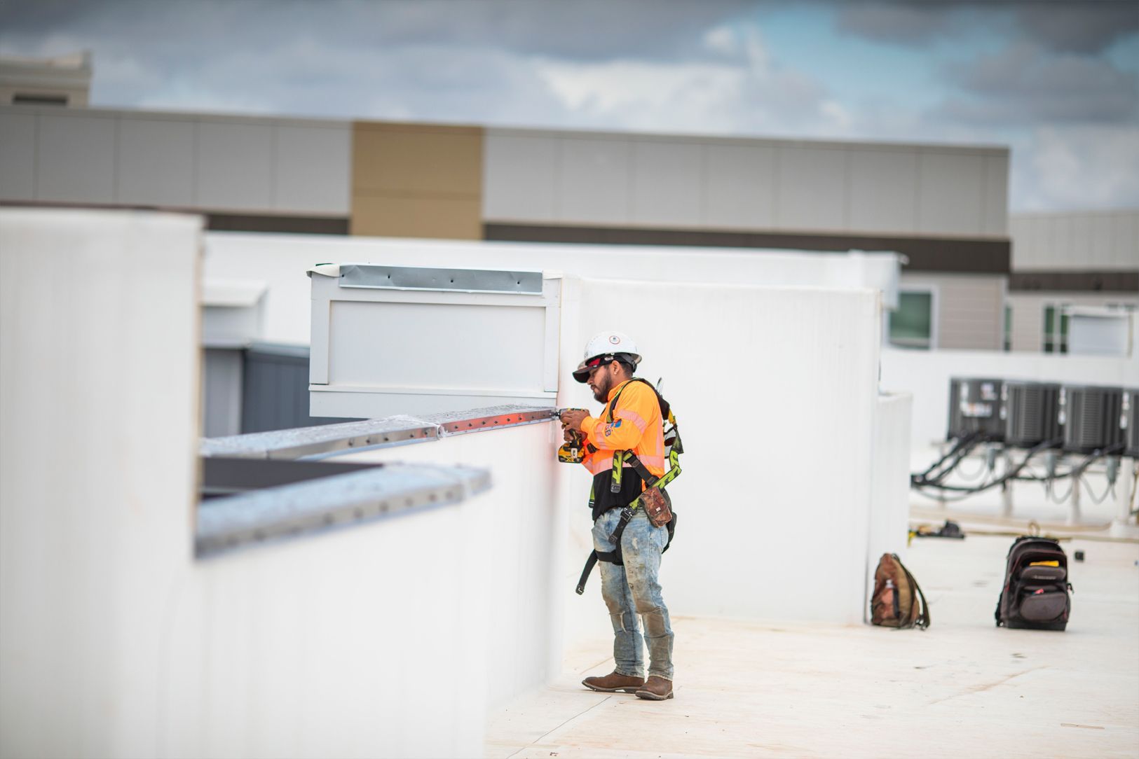 Worker in orange safety gear using a rope on a rooftop near equipment and backpacks