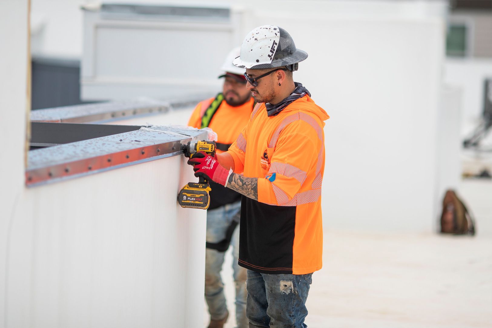 Two workers in orange safety gear measuring a rooftop edge with a tool