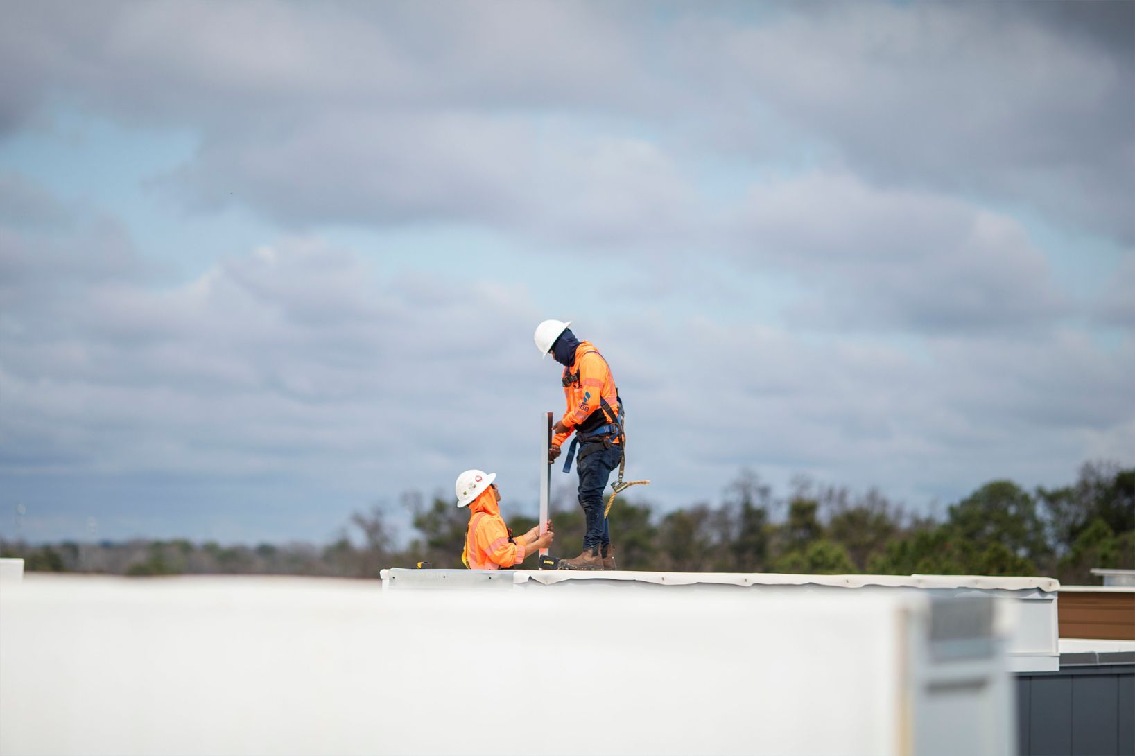 Two workers in orange safety gear on a rooftop under a cloudy sky