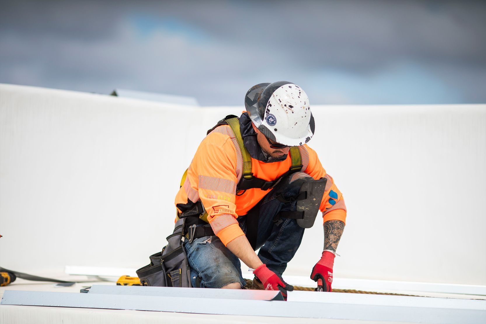 Construction worker in orange vest and white hard hat kneeling on a rooftop, measuring or installing material