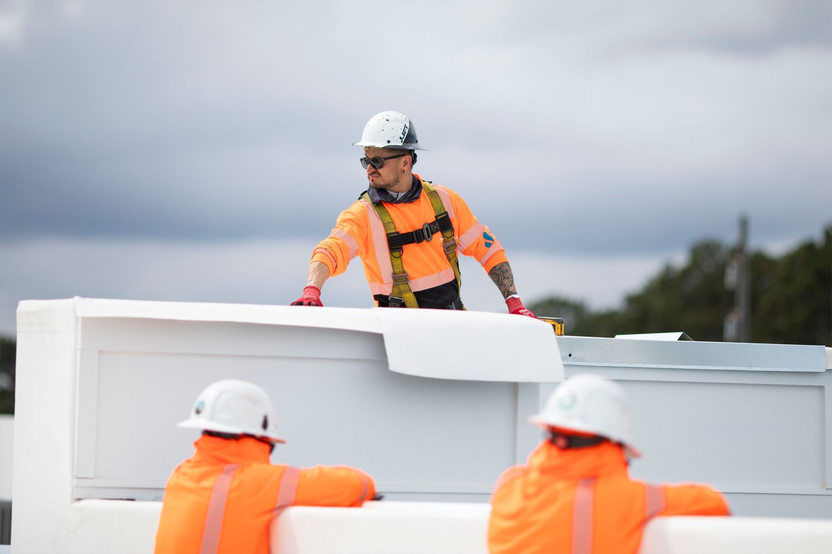 Construction workers in orange safety gear lifting a white panel outdoors under cloudy skies