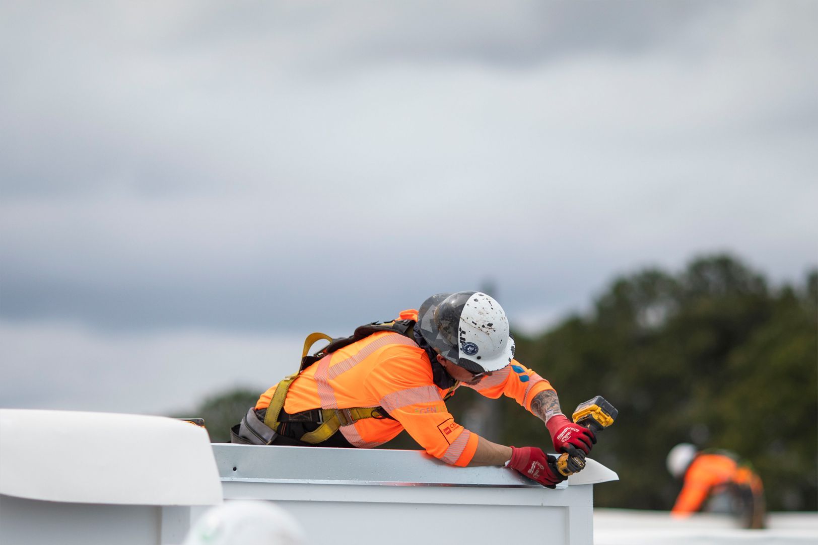 Worker in orange safety gear kneeling on a rooftop, securing something near the edge under a cloudy sky