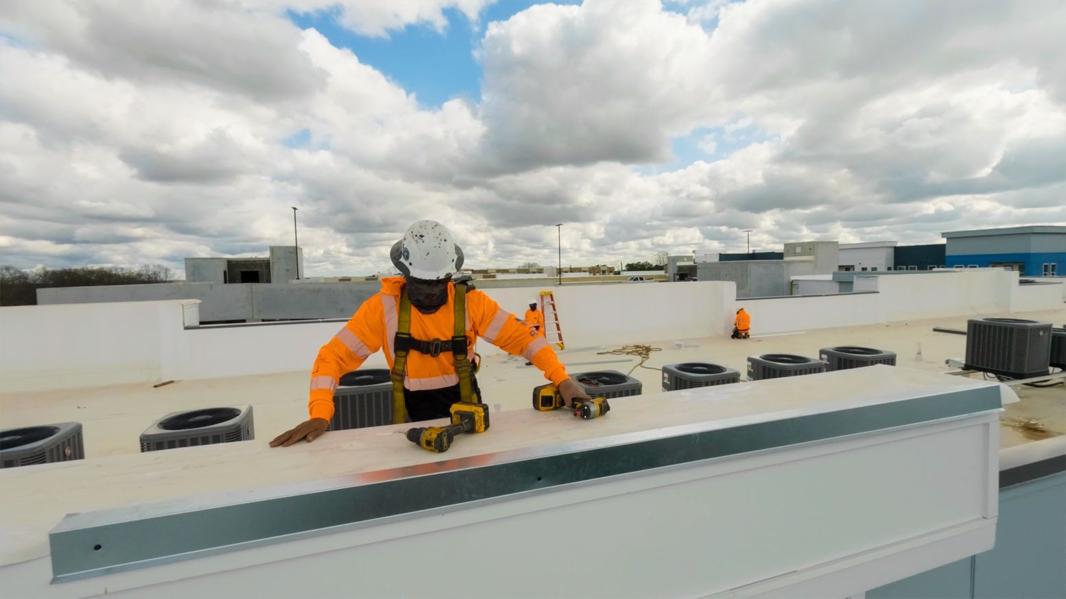 Worker in orange safety gear kneeling on a rooftop, inspecting equipment under a cloudy sky.