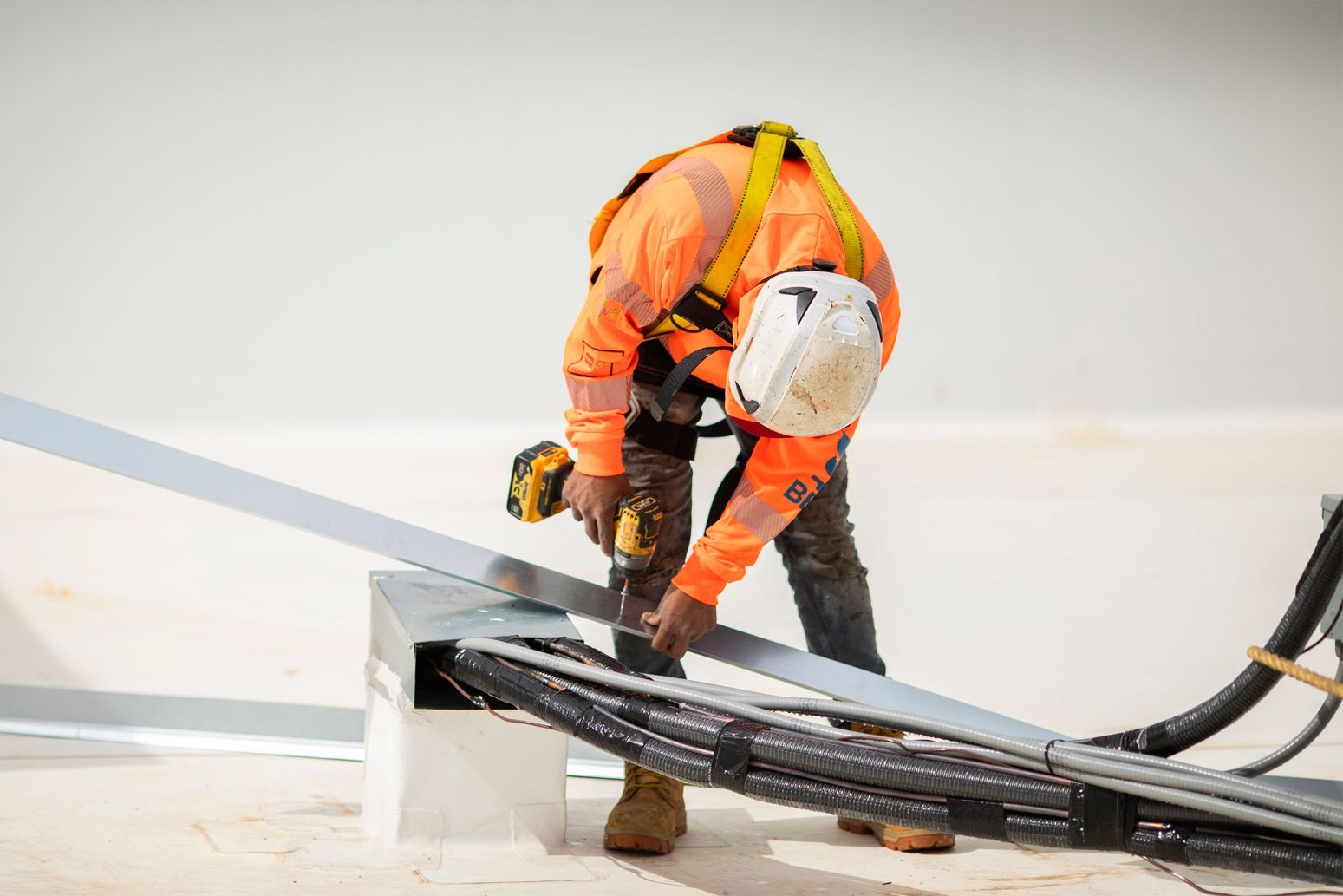 Worker in orange safety gear bending over cables on a white rooftop or industrial surface