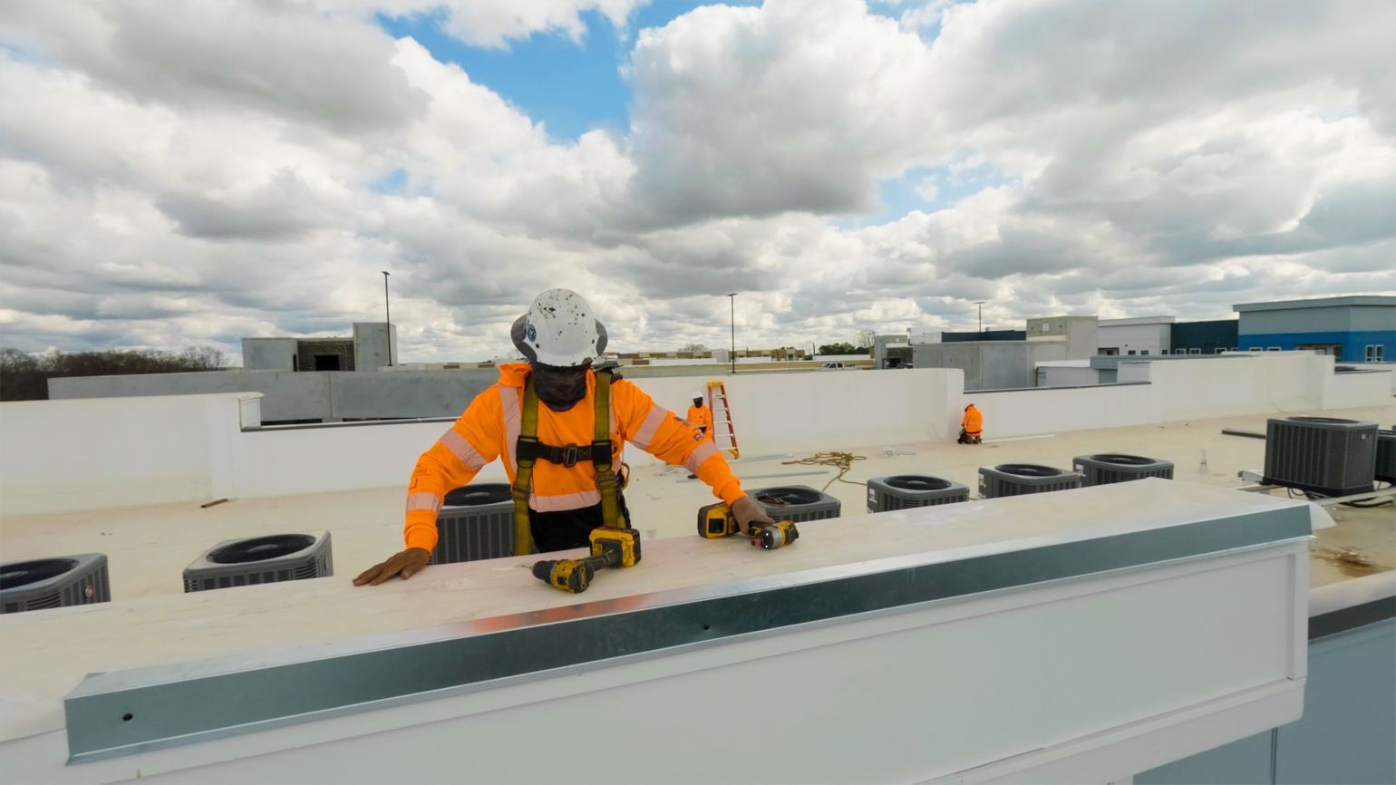 Worker in orange safety gear on a rooftop, inspecting a white roof edge under cloudy skies