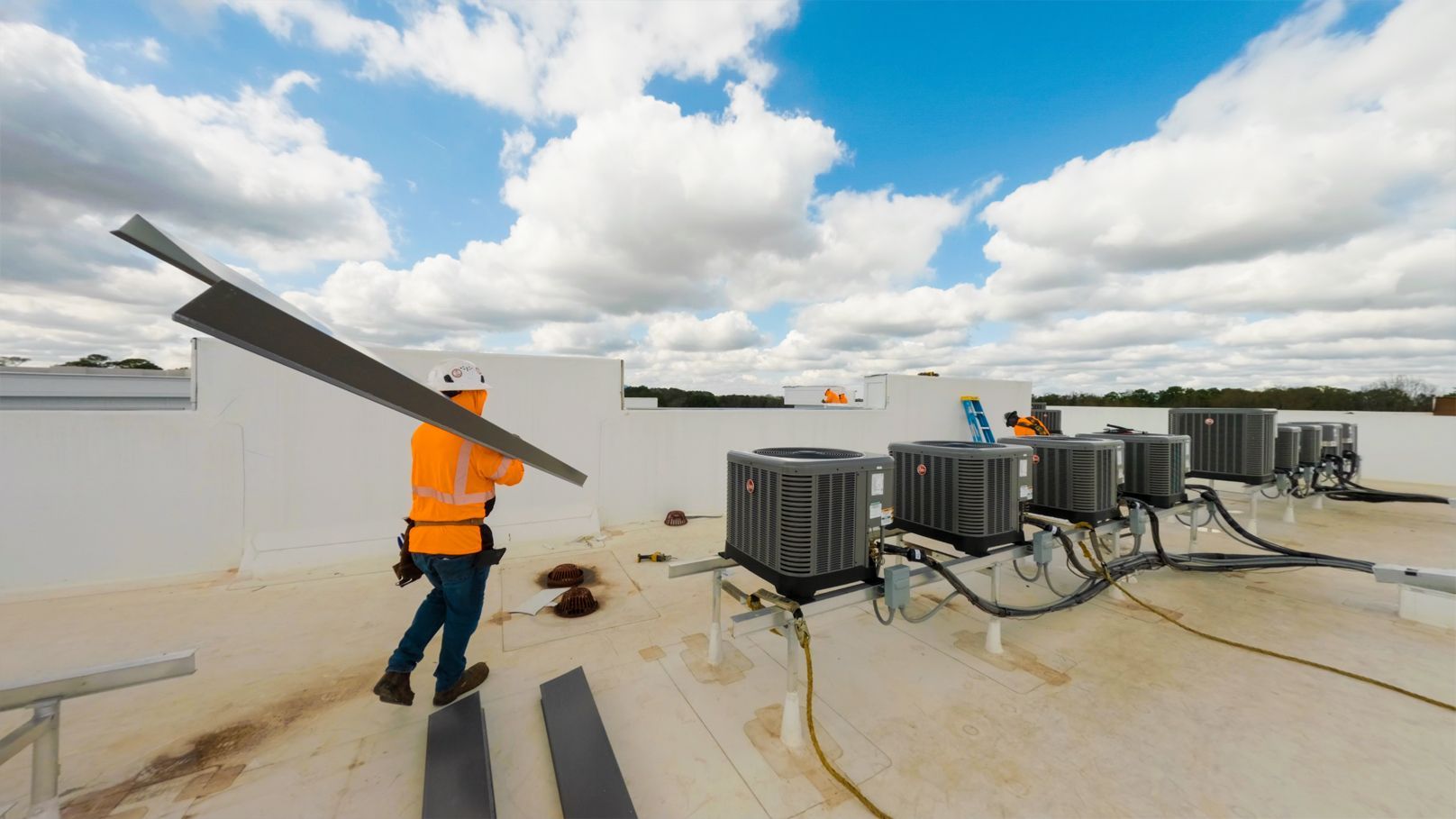 Worker in orange safety vest carrying a solar panel on a rooftop near HVAC units under a cloudy sky