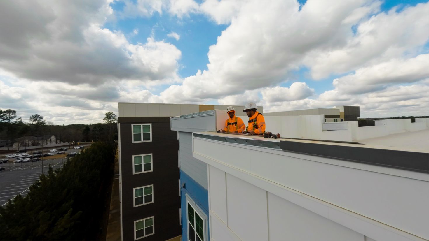Two workers in orange safety gear on a rooftop beside a white building under a cloudy sky