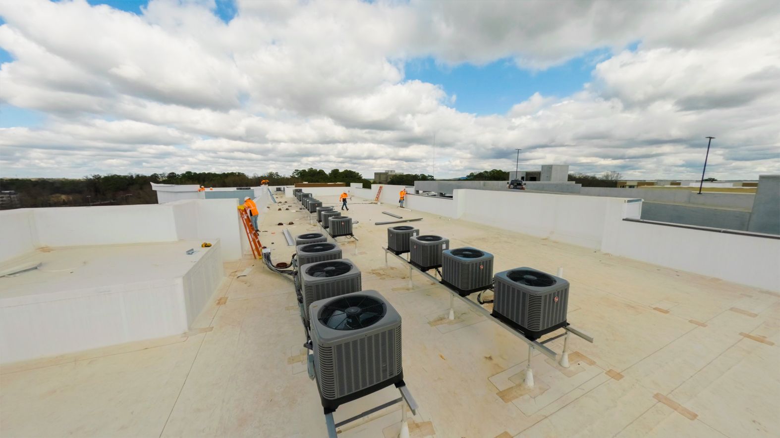 Rooftop HVAC units lined up on a flat roof under a cloudy sky, with two workers in orange vests.