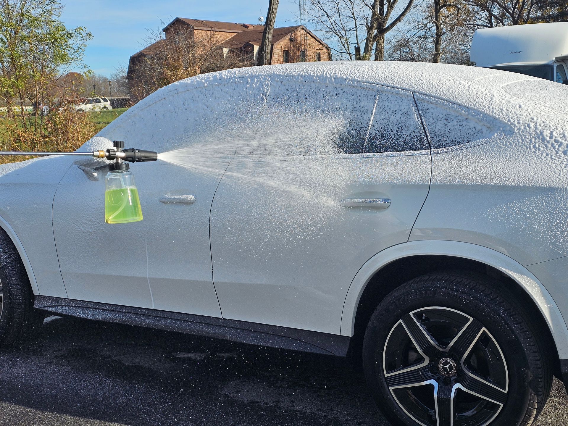 White car covered in foam being washed with a spray nozzle outdoors.