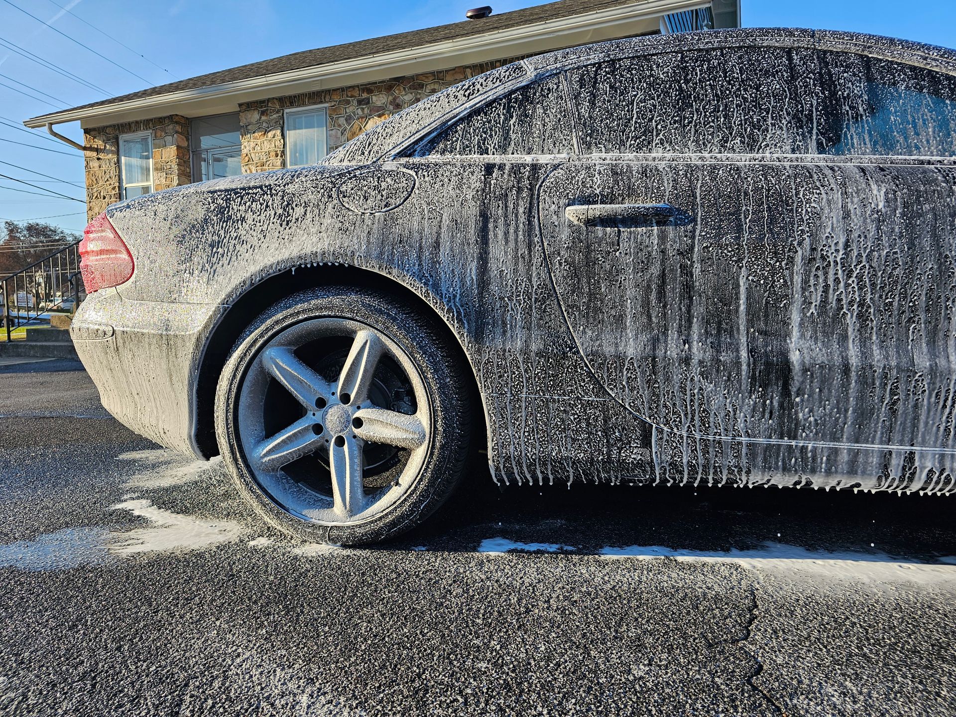Black car covered in foamy soap suds, parked on asphalt; sunlight.