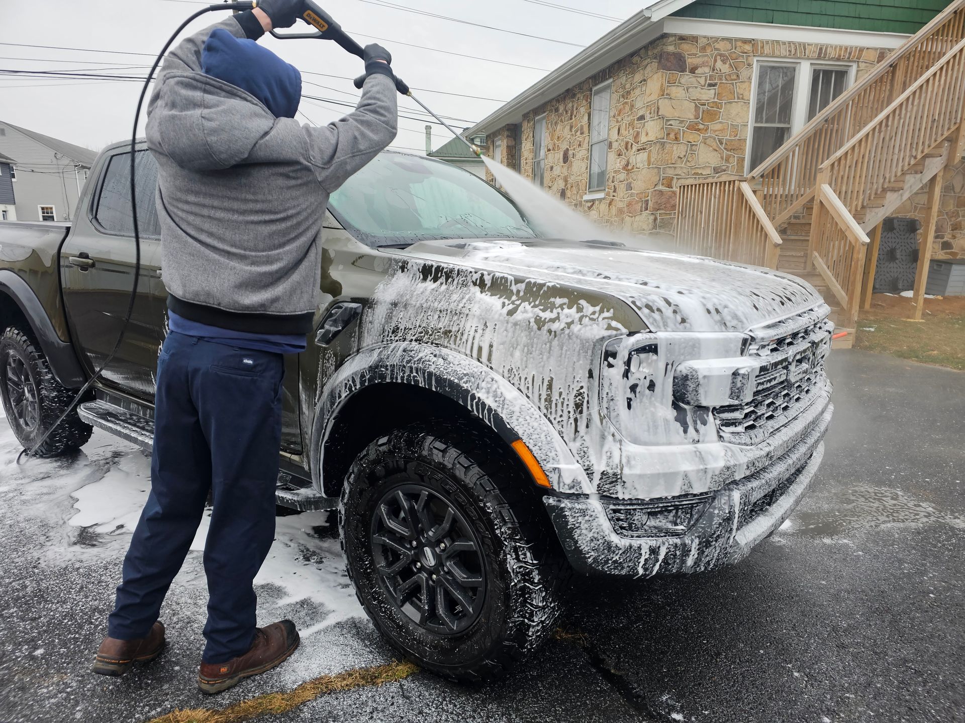 Person washing a green pickup truck with a pressure washer, covered in soap, outdoors.
