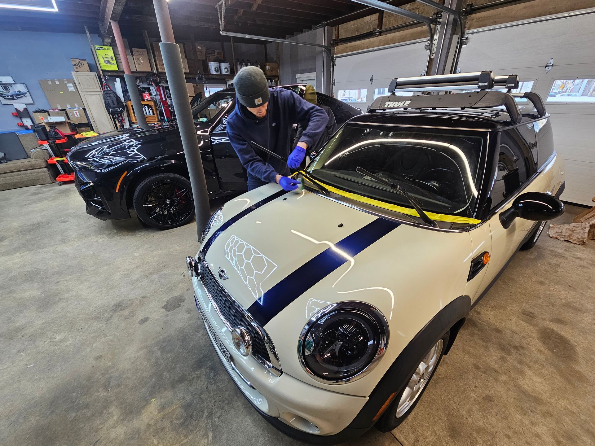 A person in a garage works on a white Mini Cooper. A black car is visible in the background.