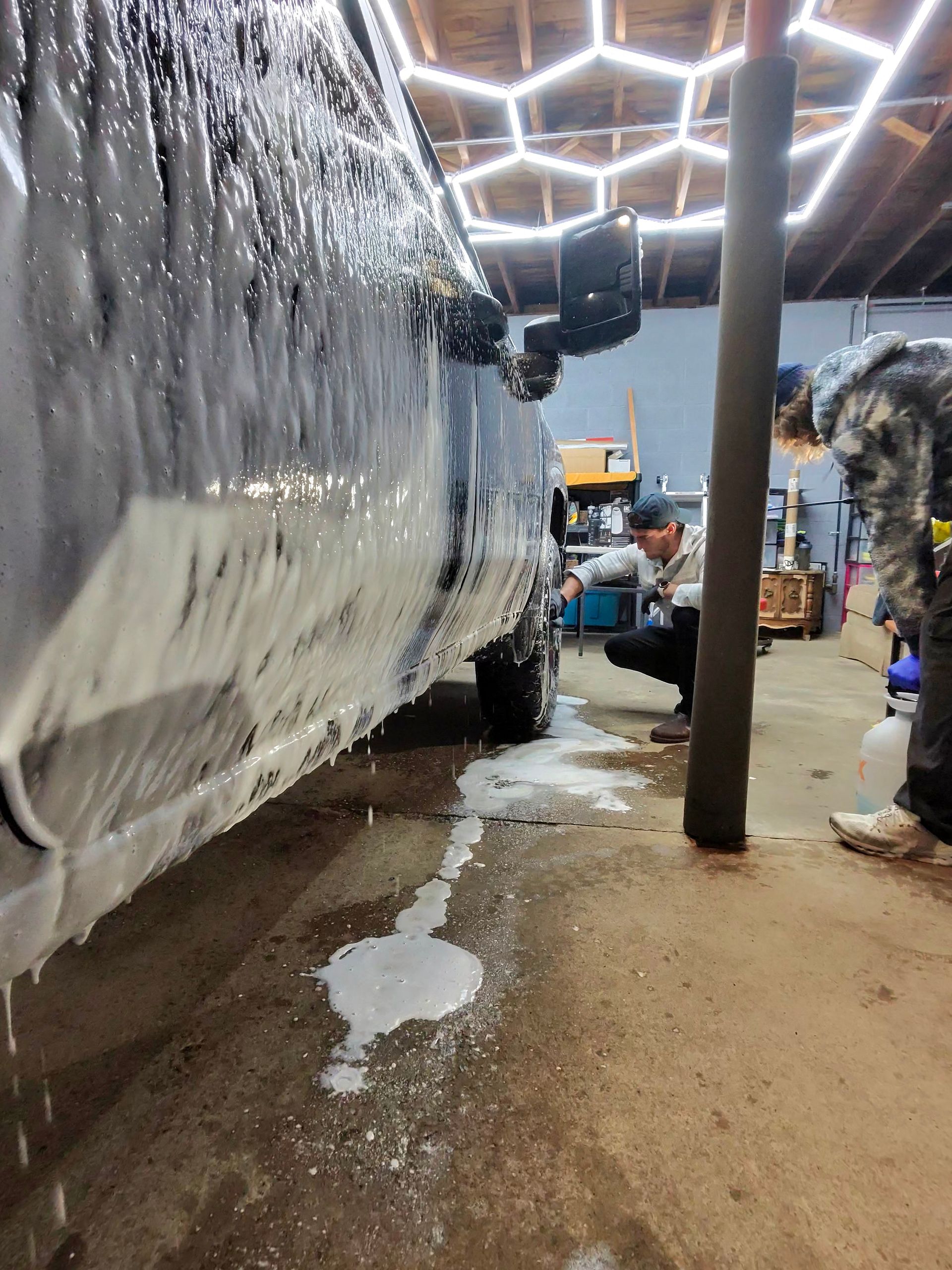 Car being washed with foam by two people in a garage under hexagonal lights.