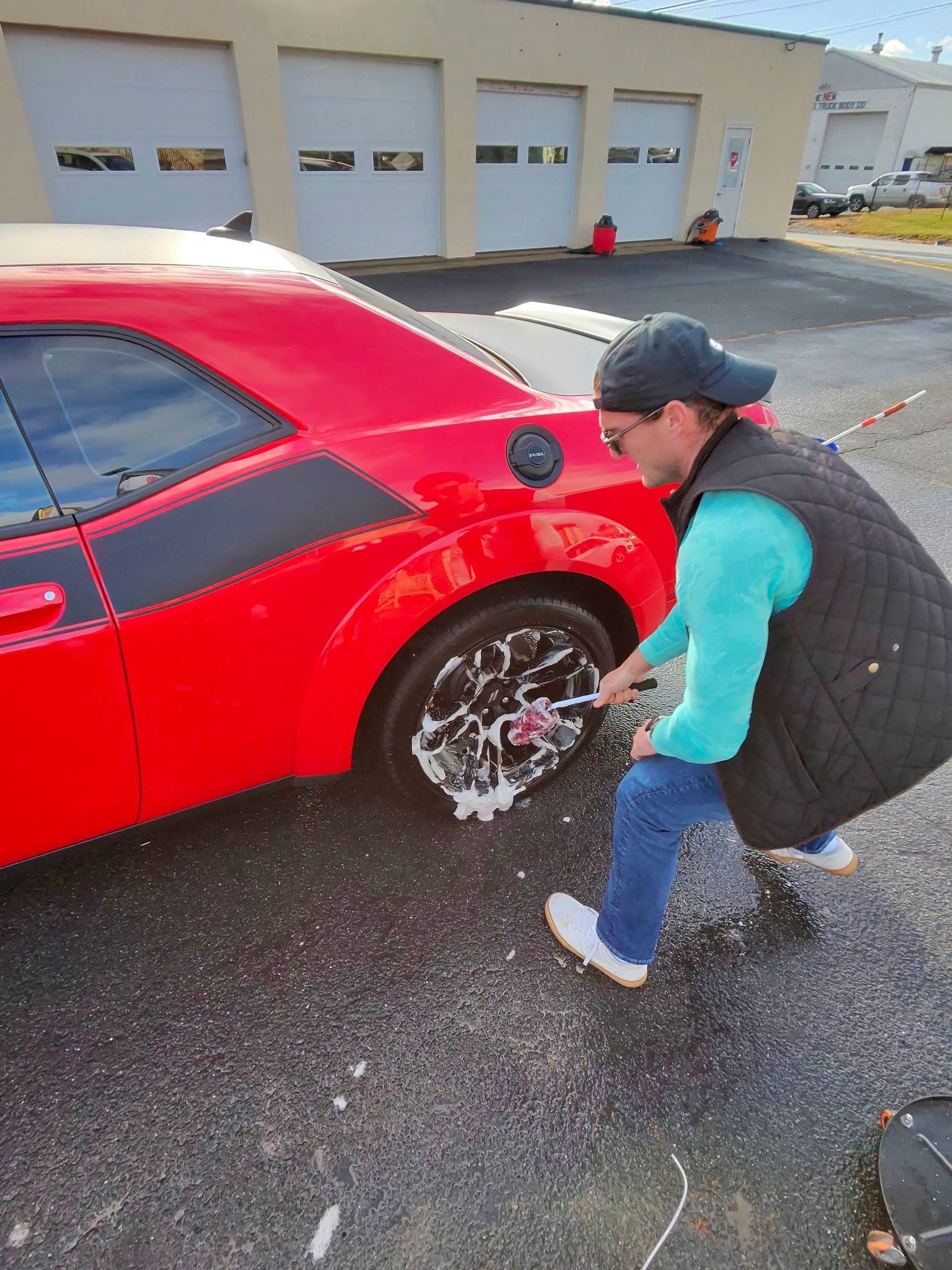 Person washing the tire of a red Dodge Challenger with soap and a brush outdoors.