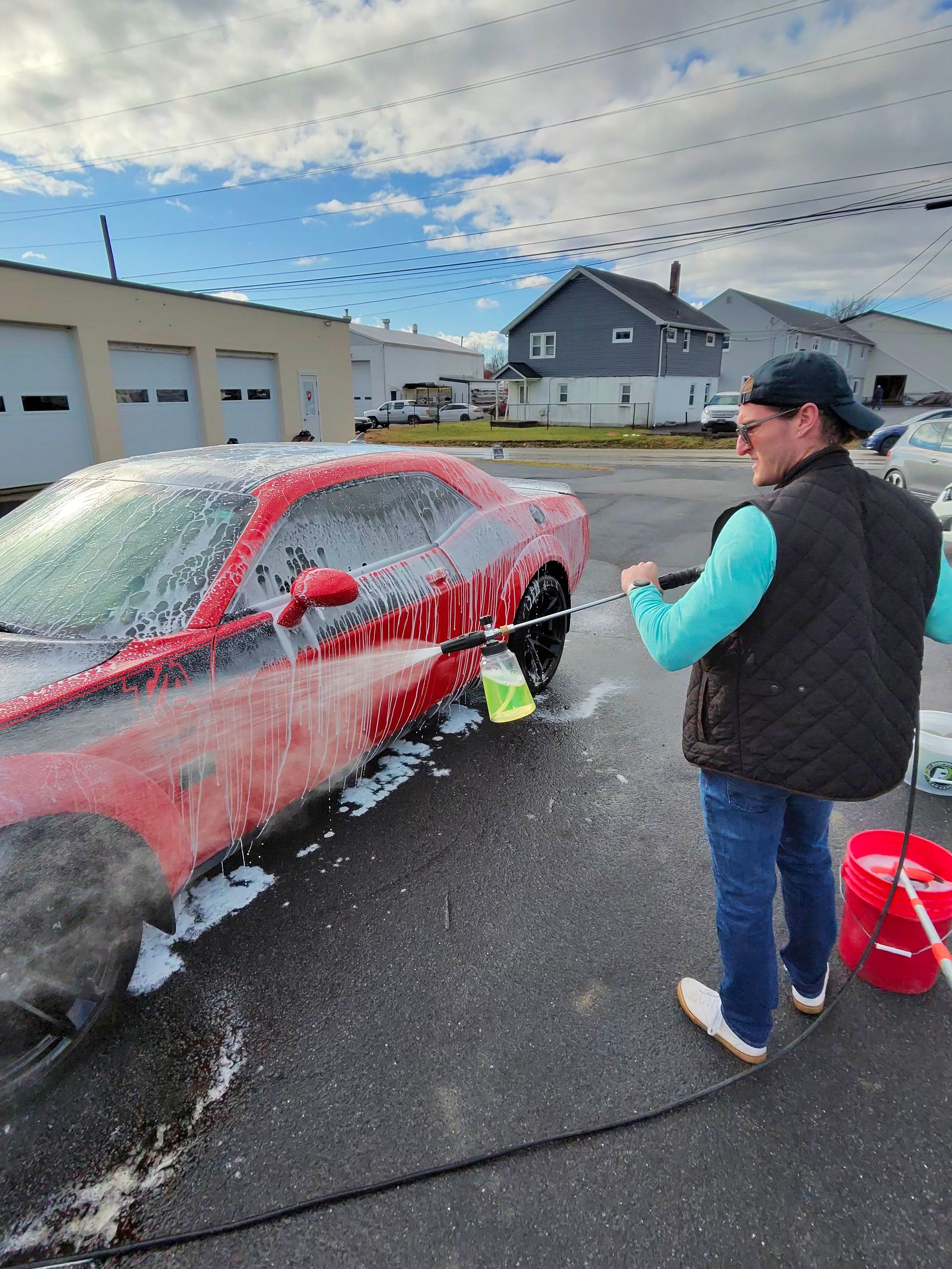 Person washing a red car with a pressure washer; outdoors on a sunny day.