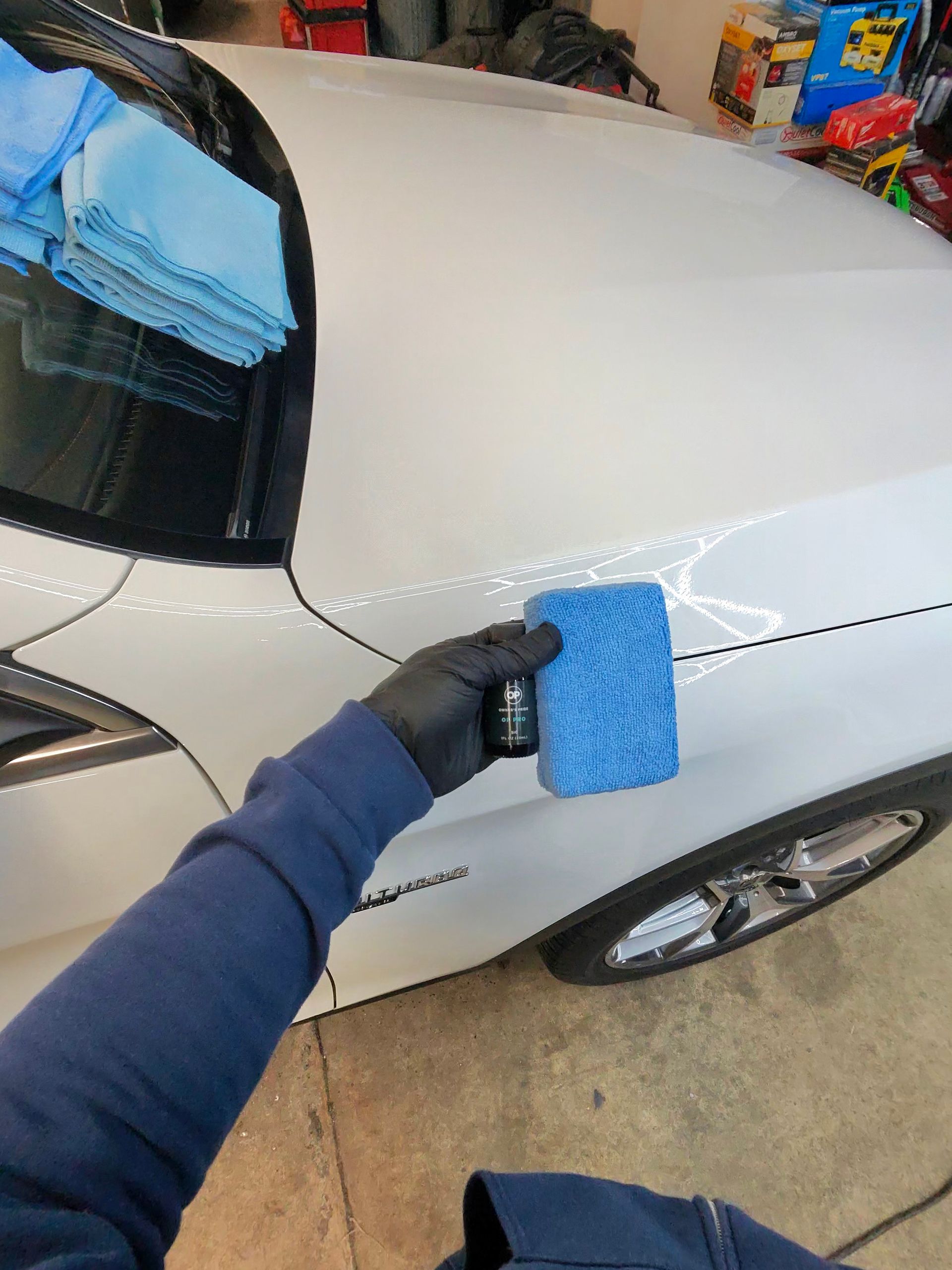 Person wearing a glove applying a blue pad to a white car's fender. Blue microfiber towels sit on the windshield.