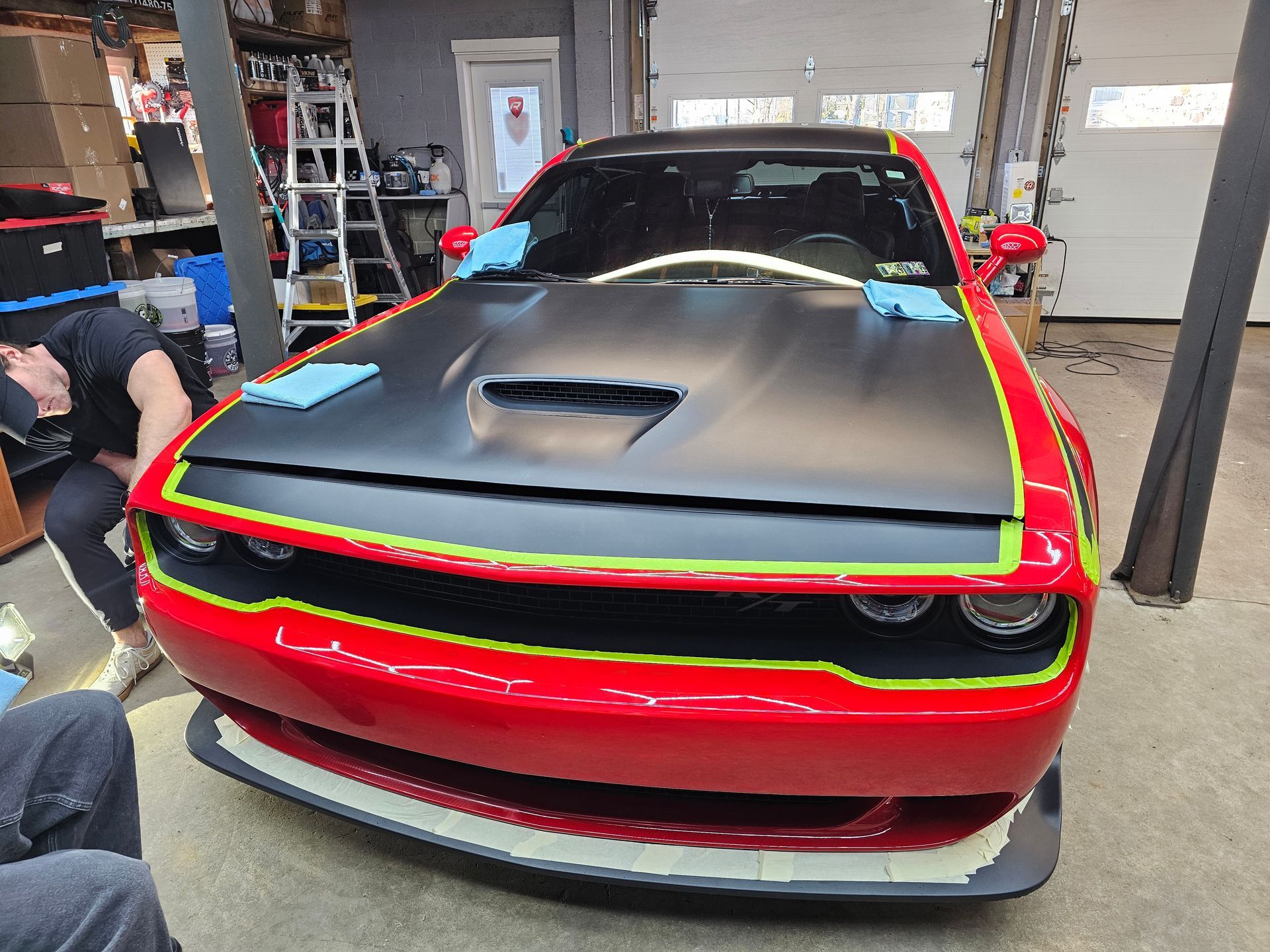 Red Dodge Challenger being worked on by a person in a garage; hood is black, with yellow and white trim.
