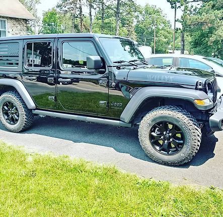 Black Jeep Wrangler Willys parked on a paved surface next to green grass on a sunny day.