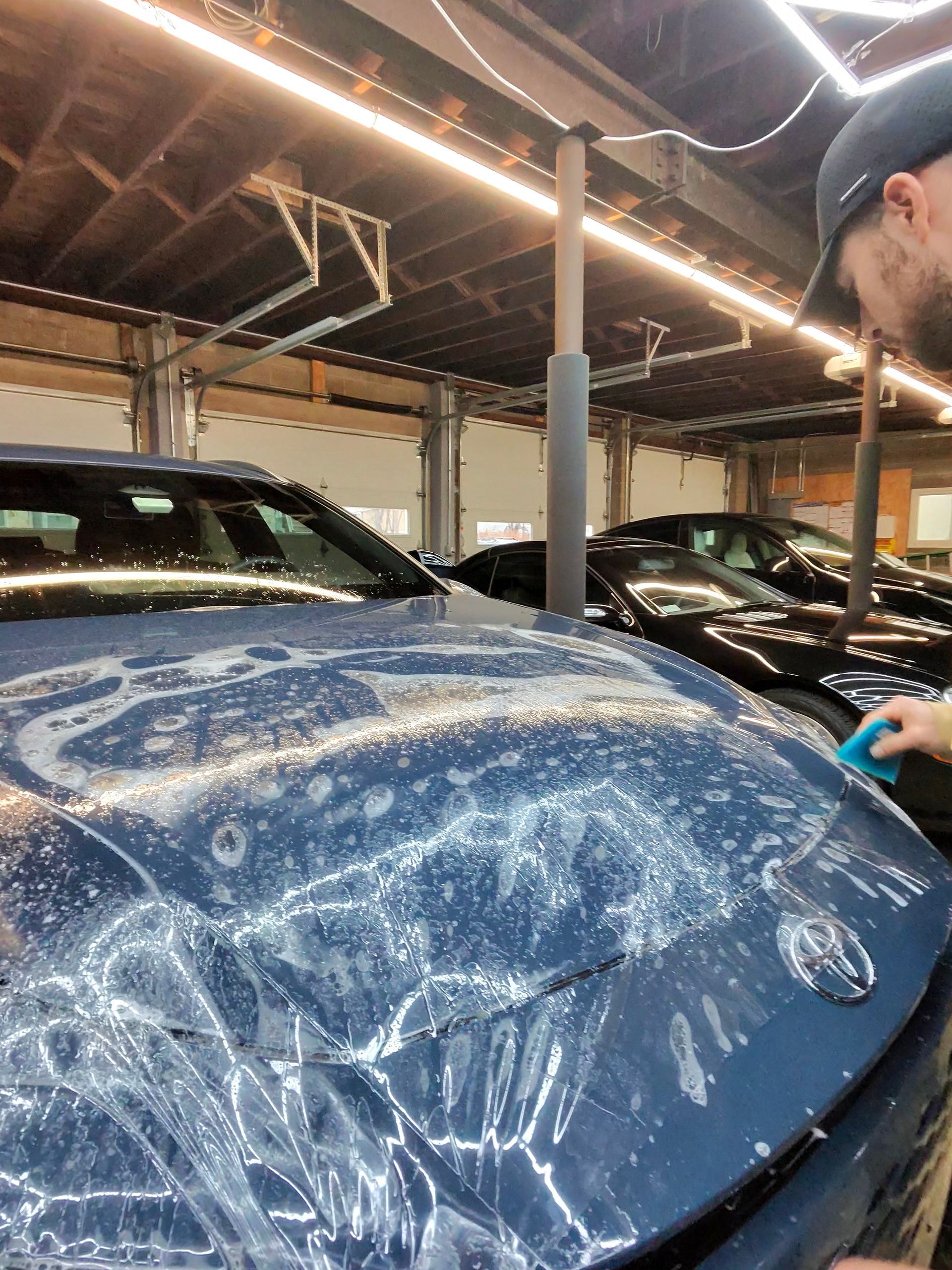 A person applying clear film to the hood of a blue car inside a garage.