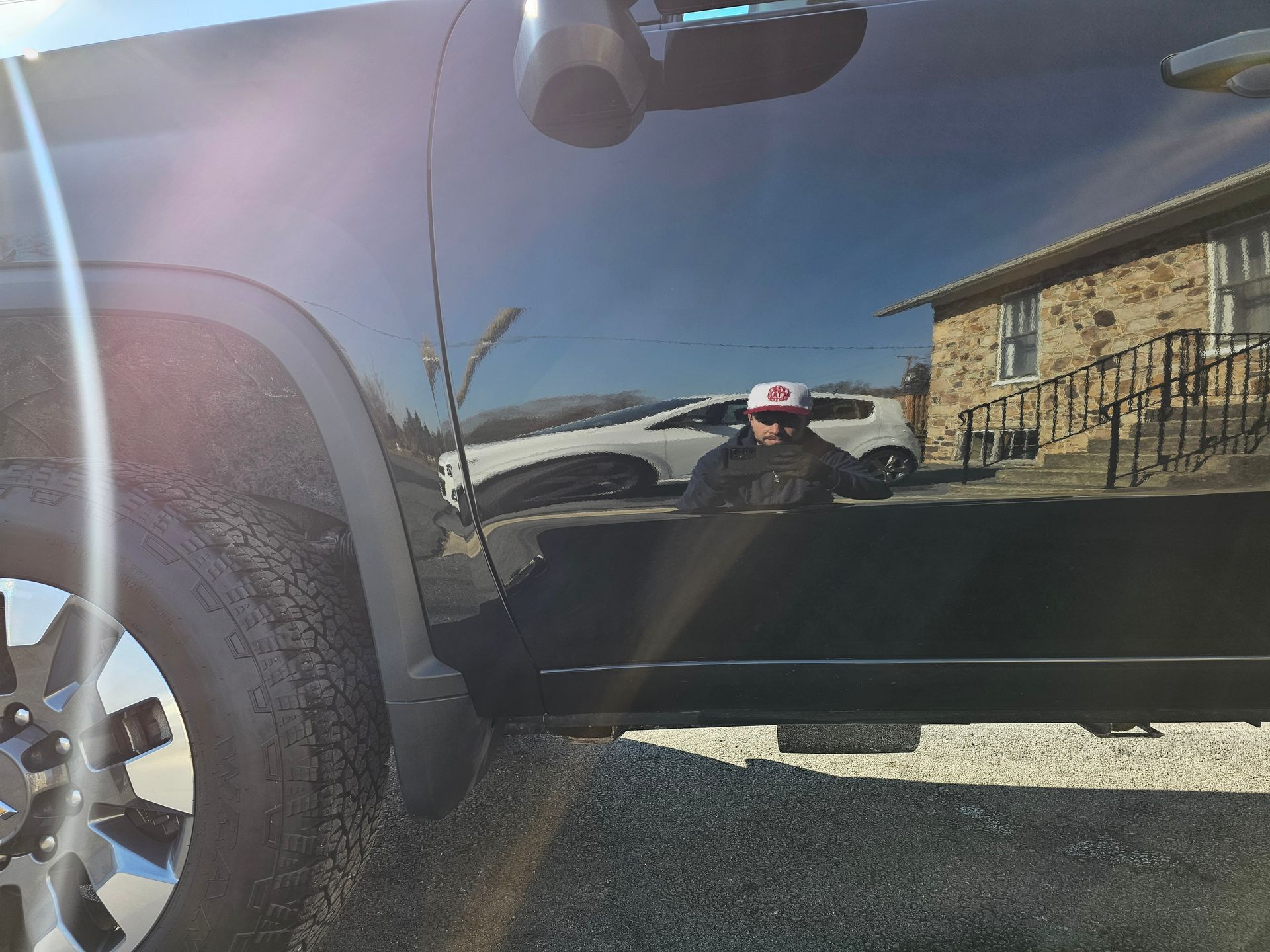 Black truck door reflecting a person in a cap, another car, and a stone building.