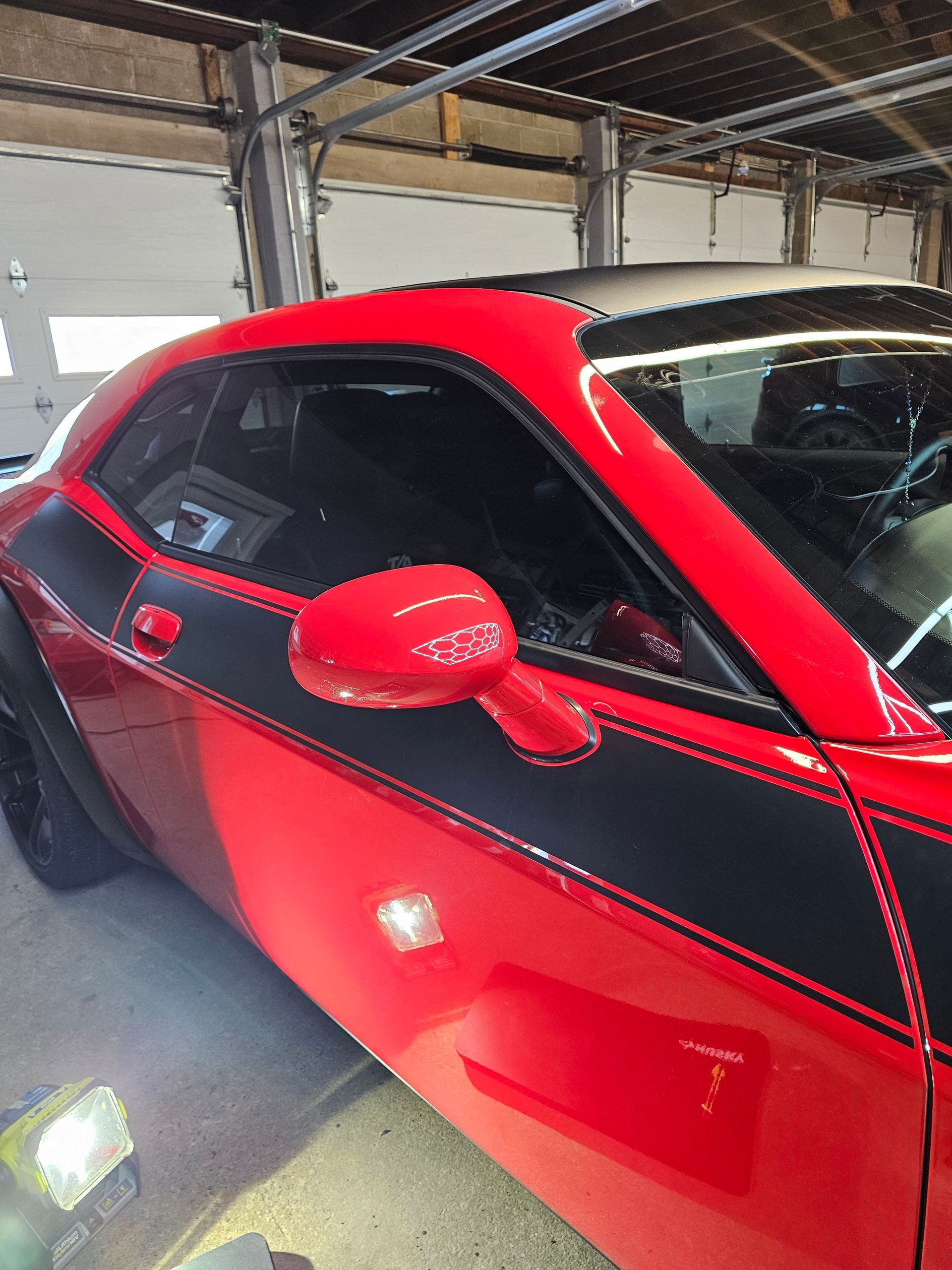 Red Dodge Challenger with black stripes, side mirror visible, parked indoors.