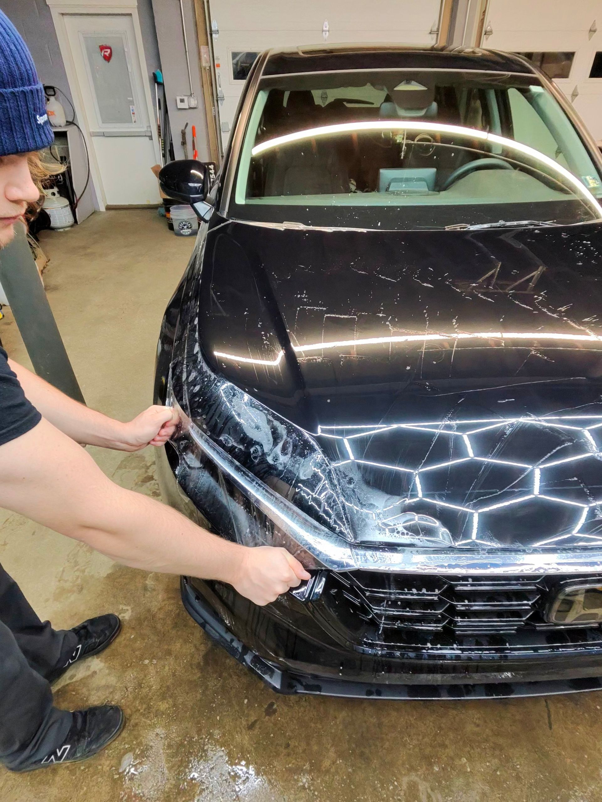 Person applying film to the front of a black car. Indoors, shop setting.