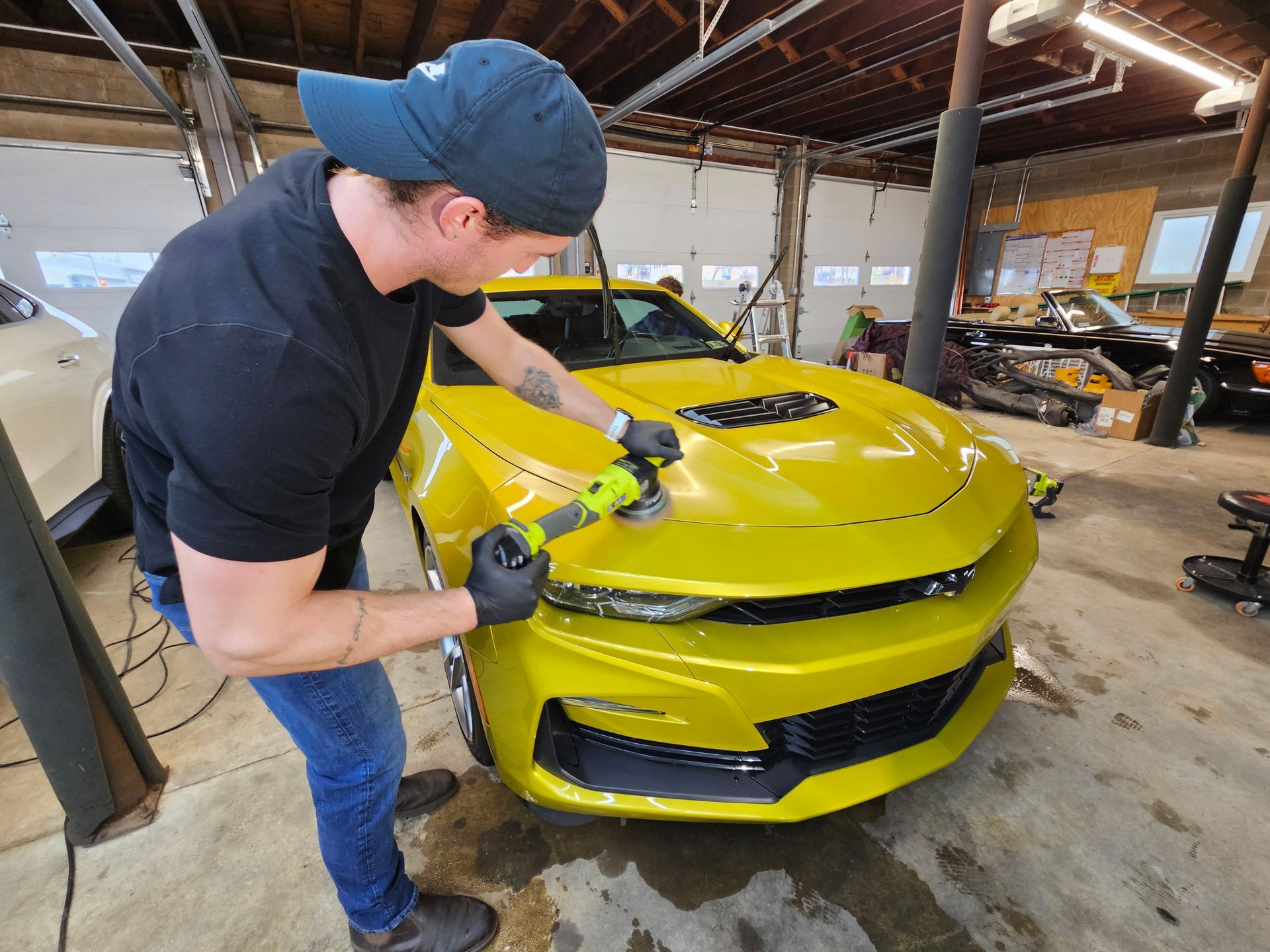 Man polishes a bright yellow car hood with a power tool in a garage.