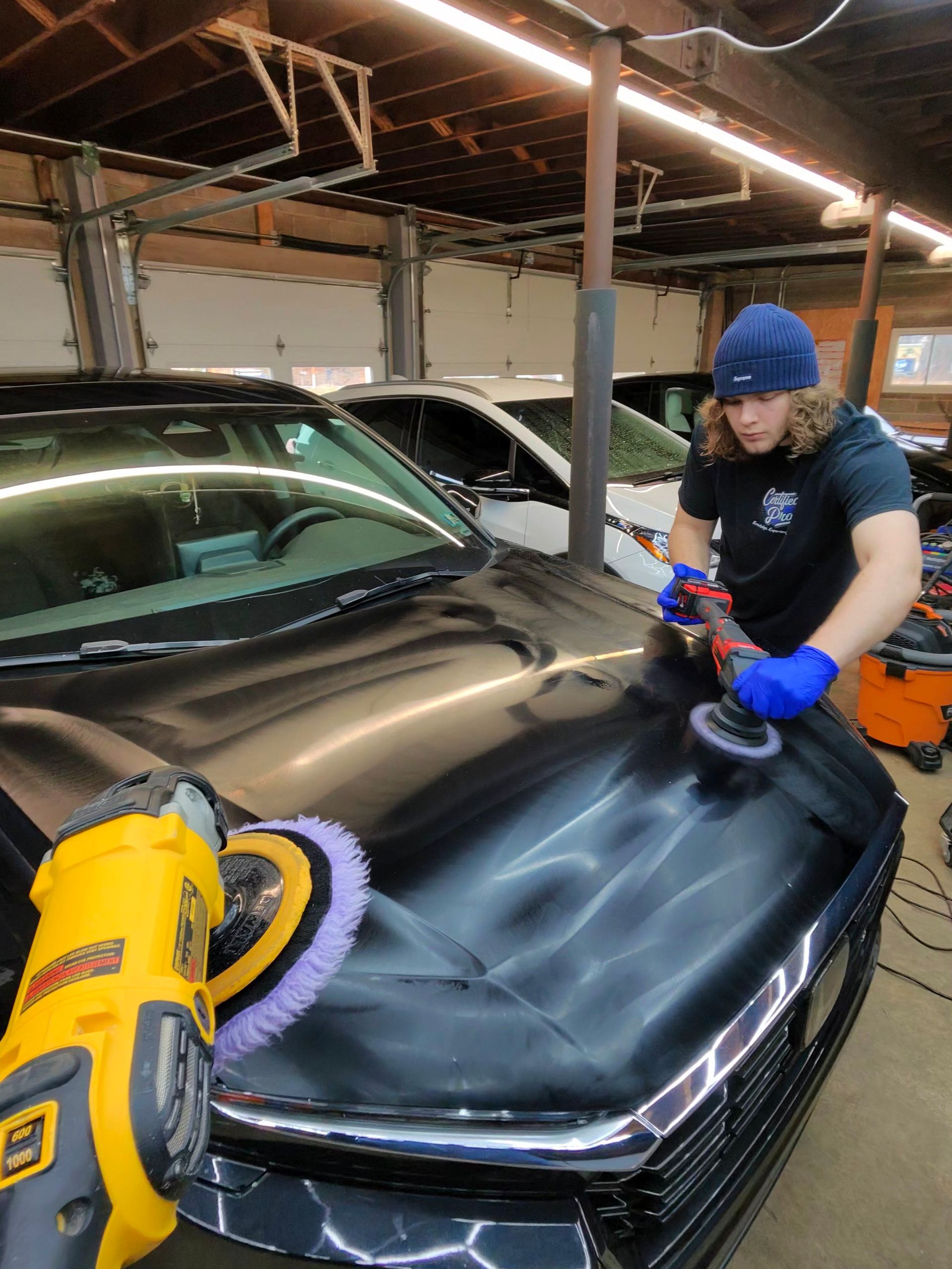 Person polishing a black car hood in a garage using an electric buffer.