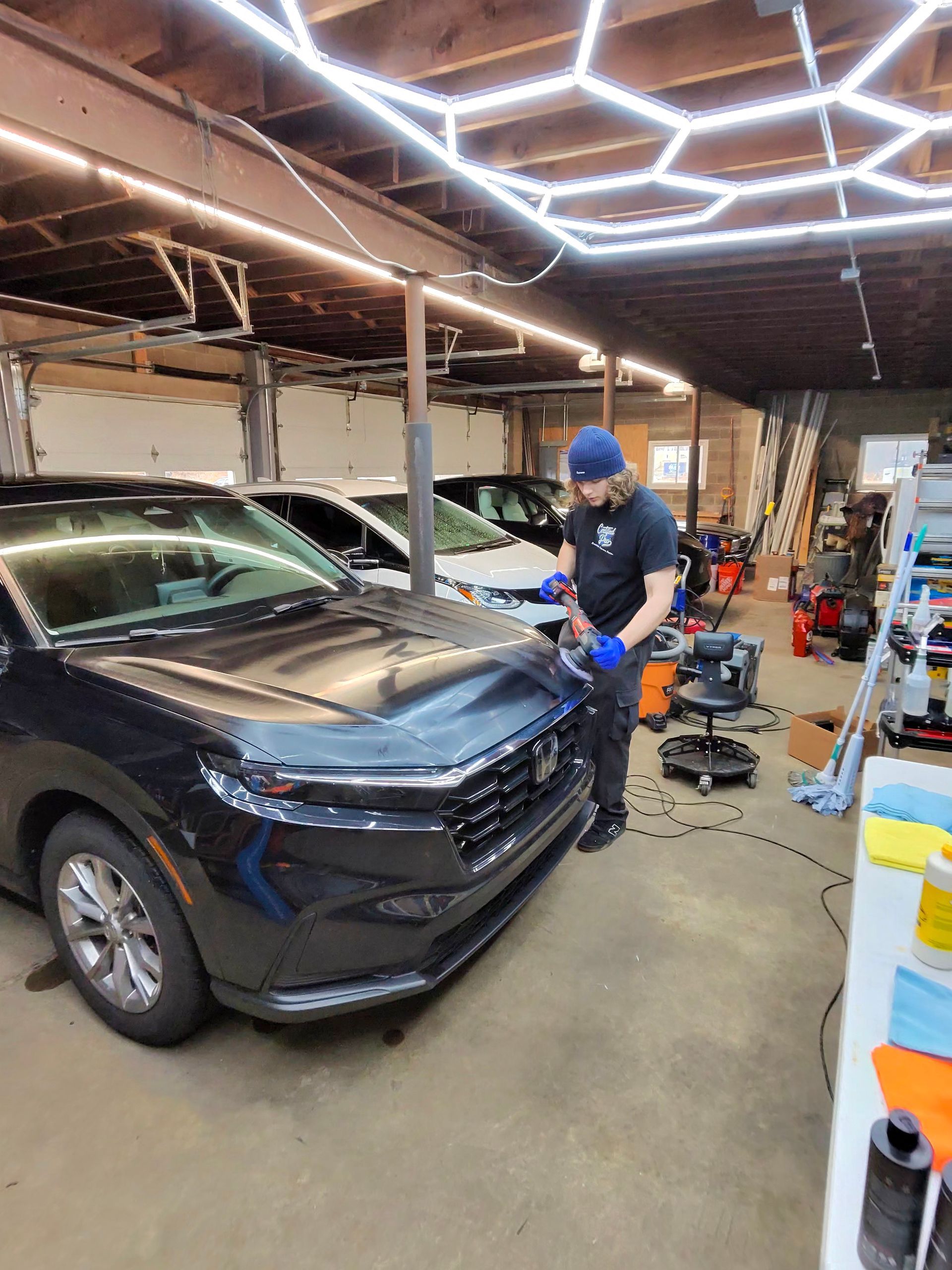 Person in garage working on a dark car; hexagon lights overhead.
