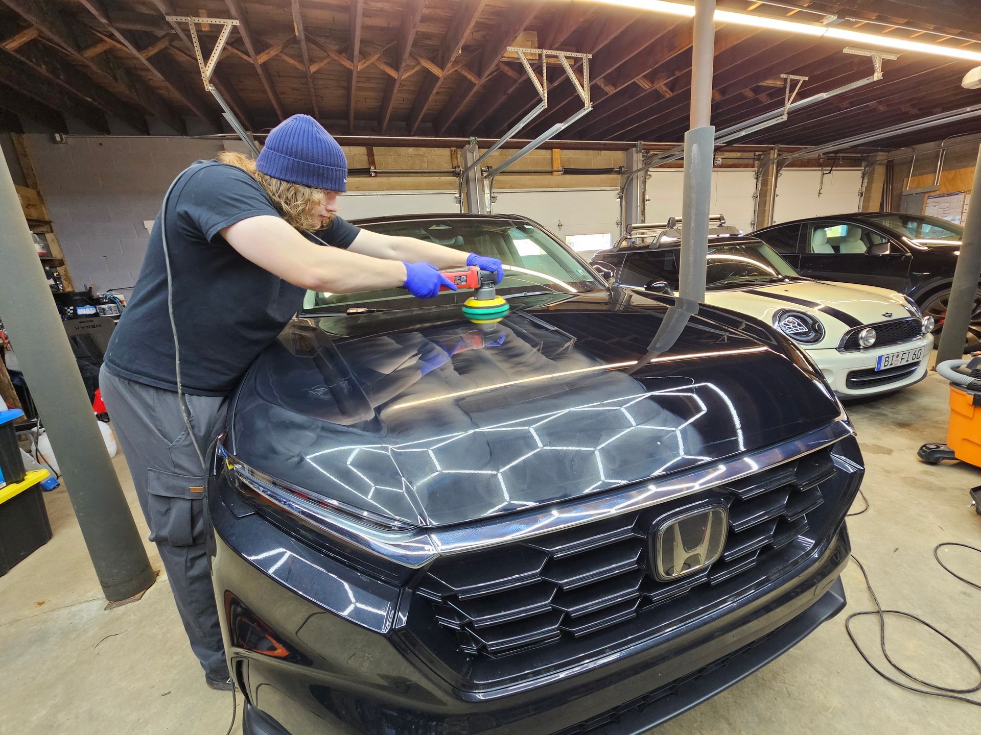 Person polishing a black car hood in a garage. Other cars are visible in the background.