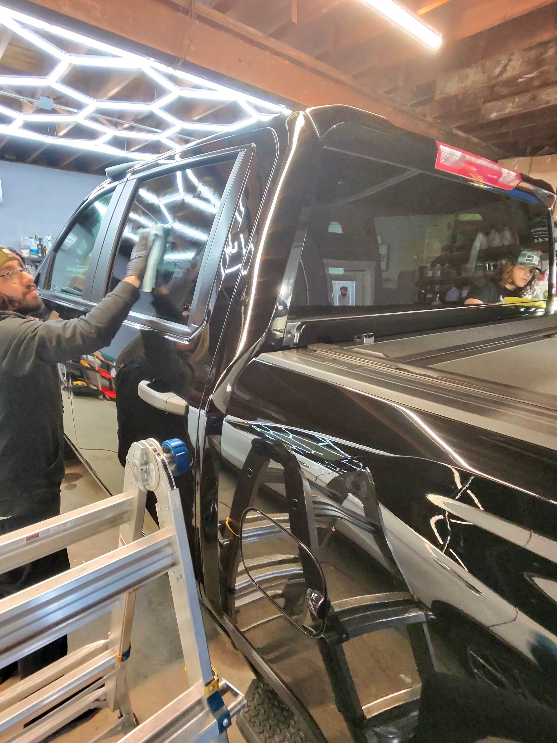 A person cleaning the window of a black truck in a brightly lit garage.