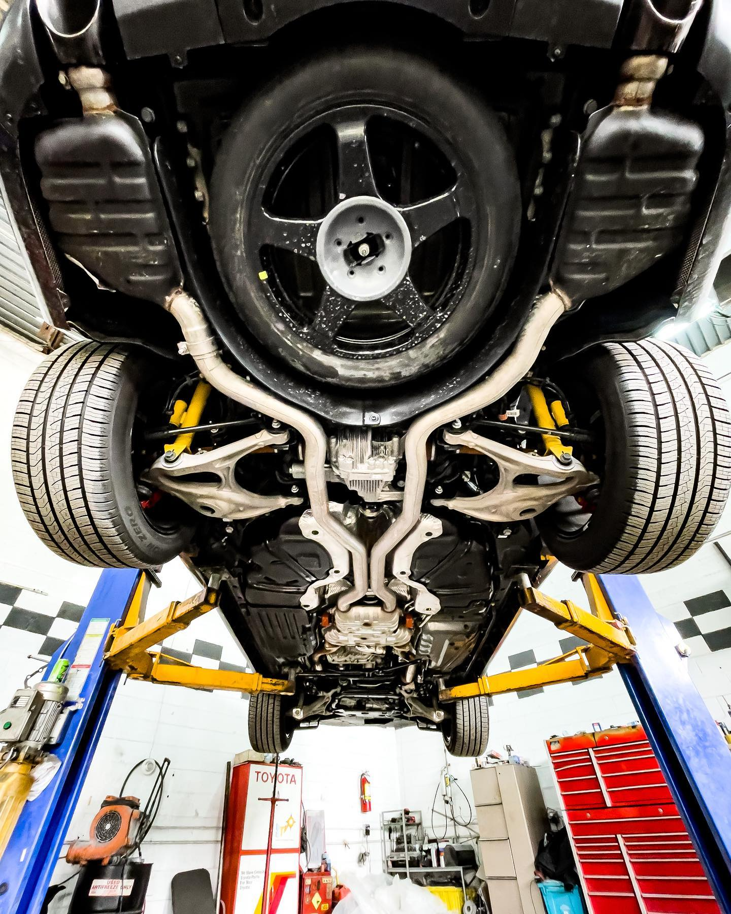 Underside view of a black car raised on a lift in a garage. Spare tire, exhaust pipes, and suspension visible.