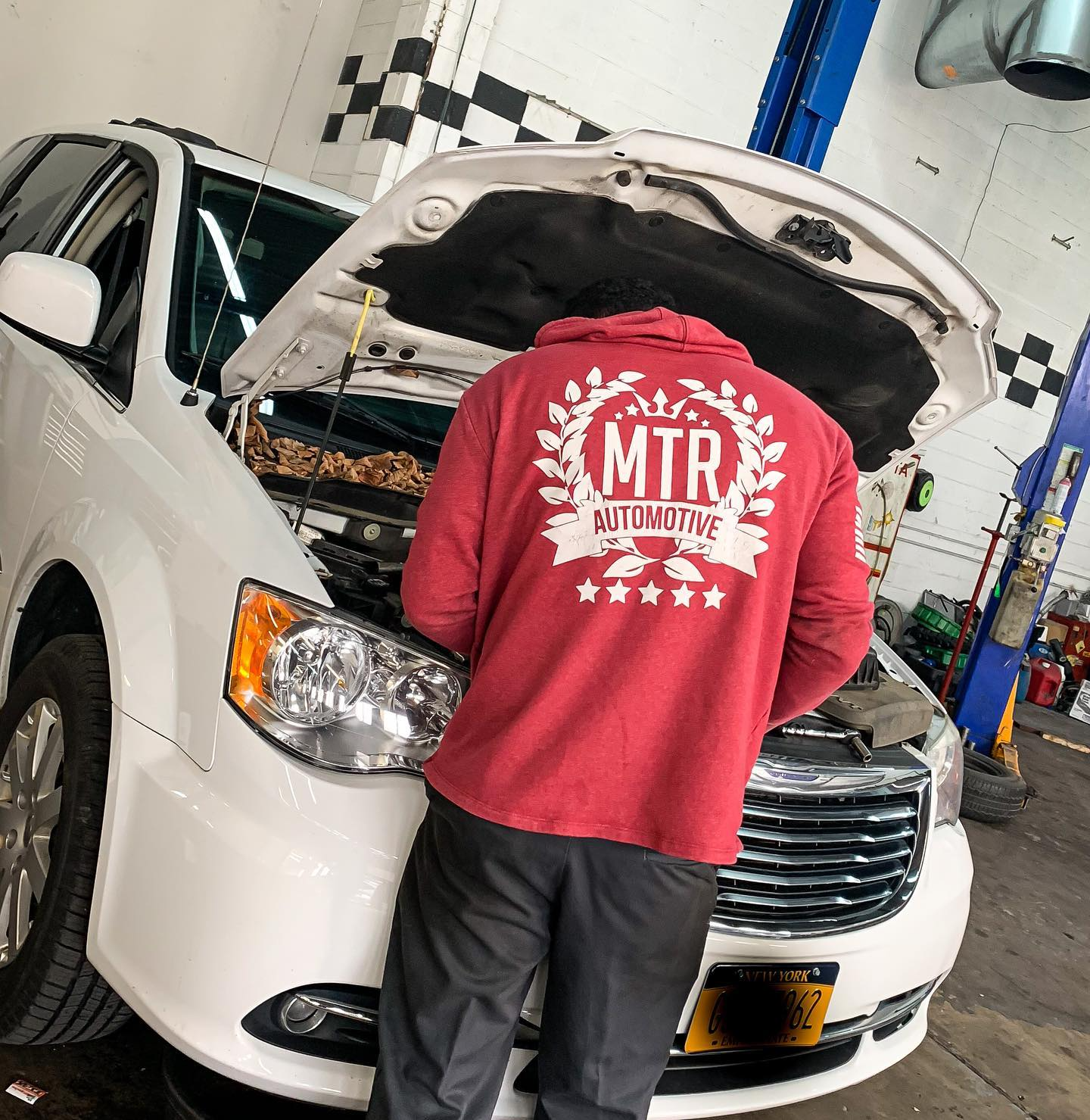 Mechanic in red hoodie inspects a white minivan in a garage.