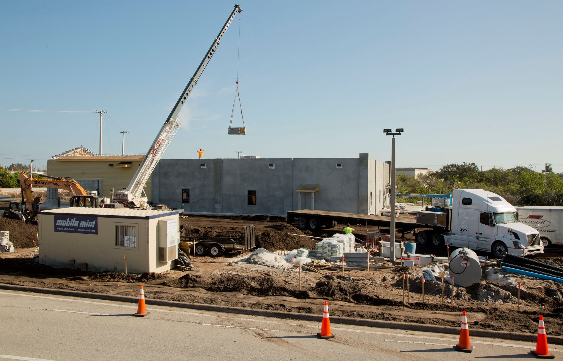 Construction site with crane lifting a wall panel beside a white truck and orange traffic cones
