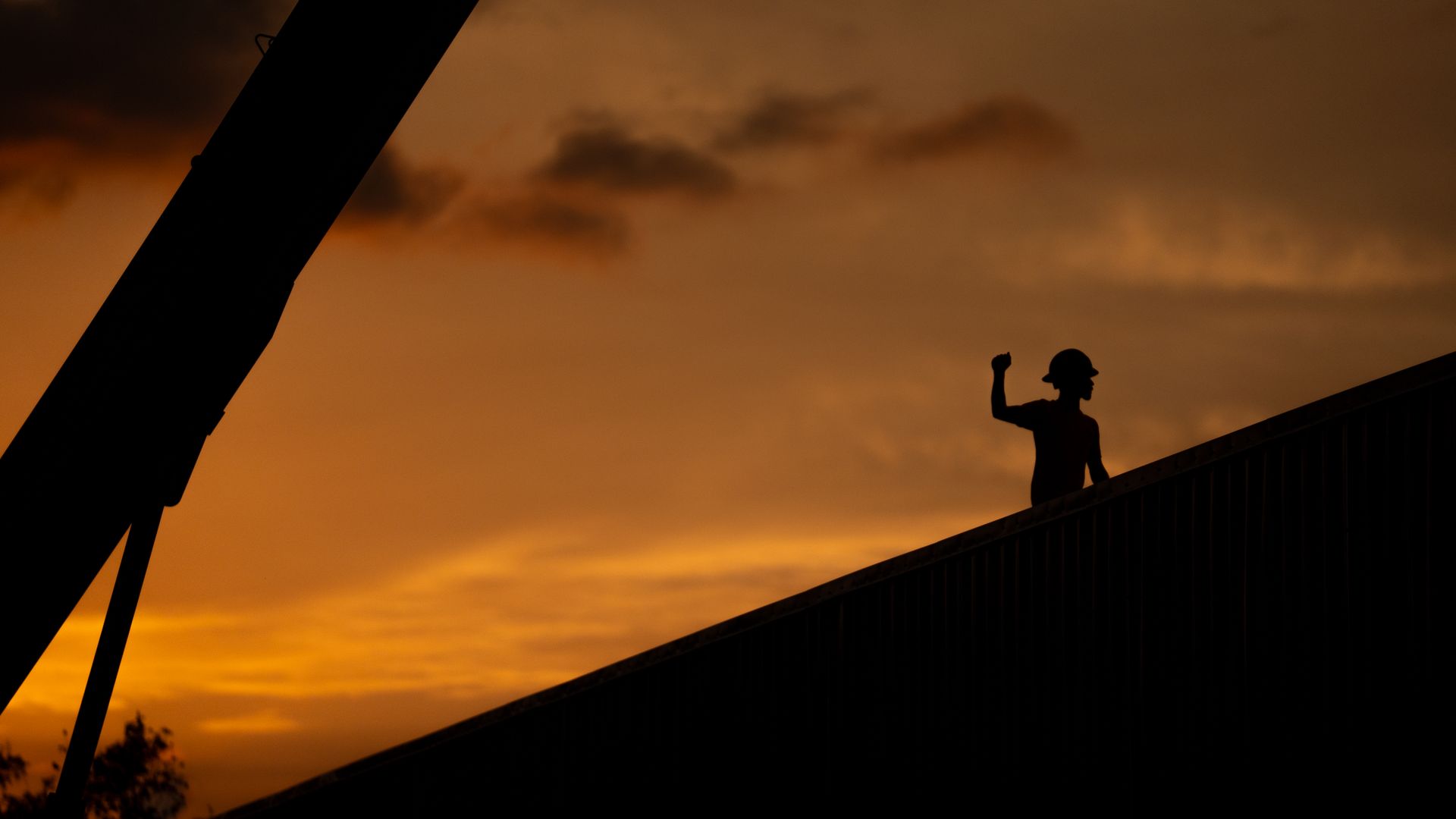 Silhouette of a person raising an arm on a rooftop at sunset, with orange sky and dark foreground