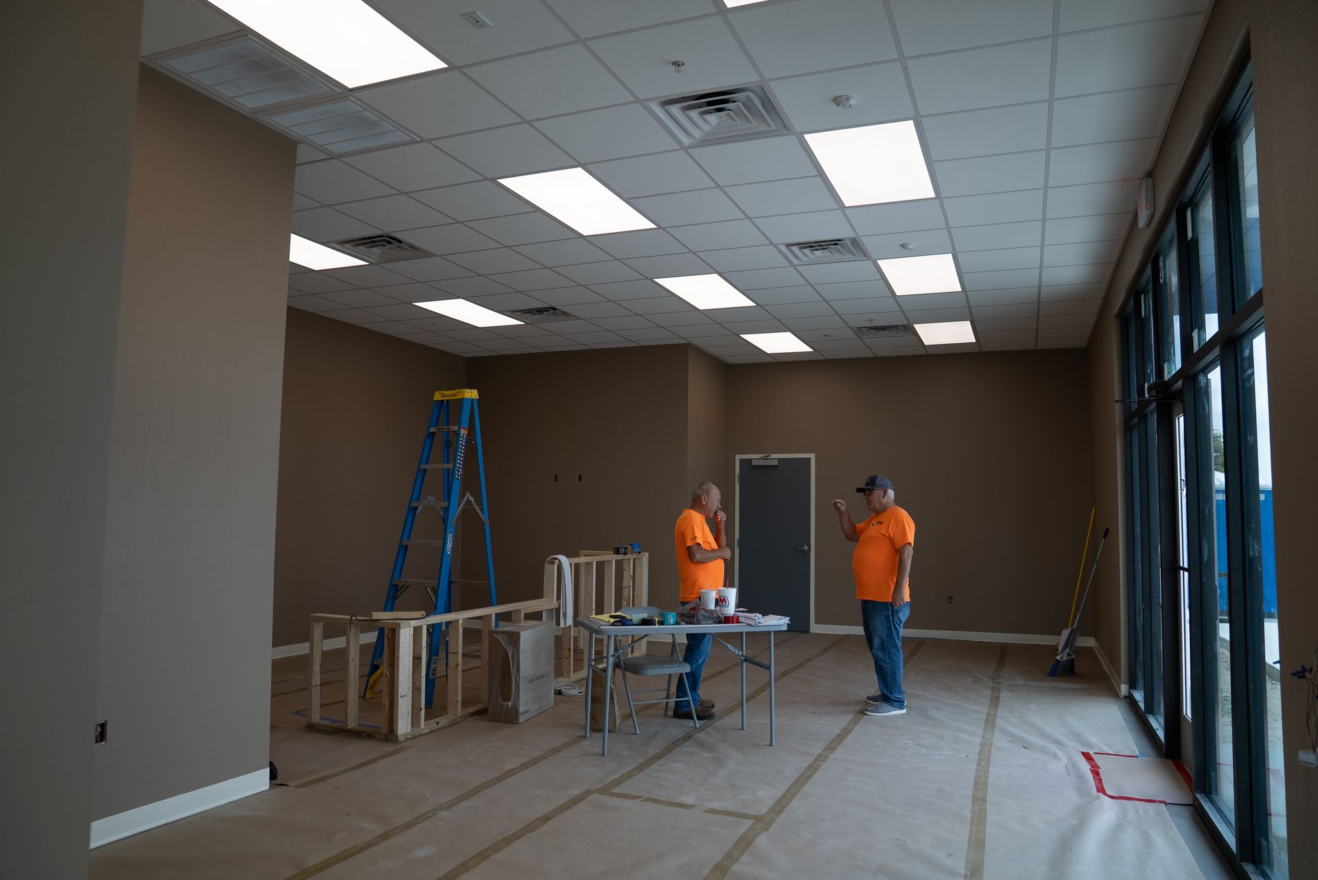 Construction workers in orange vests work in a large room with ladders and tables.