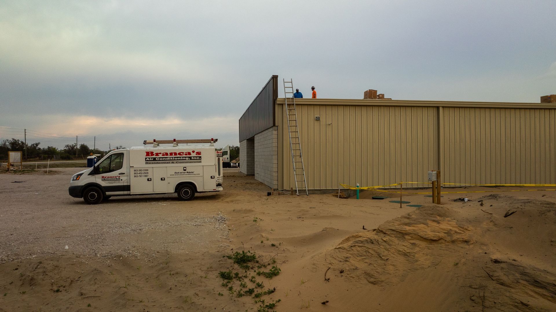 White utility van near beige industrial building in a sandy lot under an overcast sky