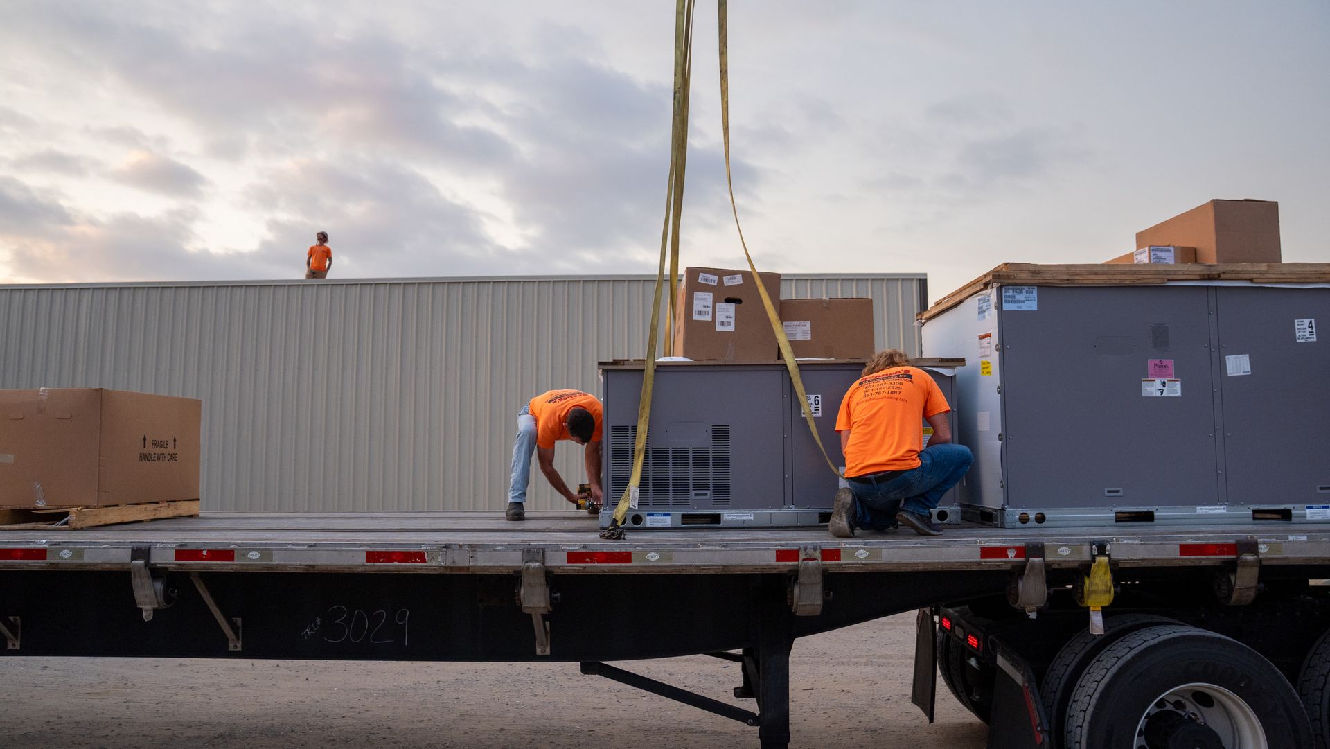 Workers in orange vests loading large crates onto a flatbed truck at dusk