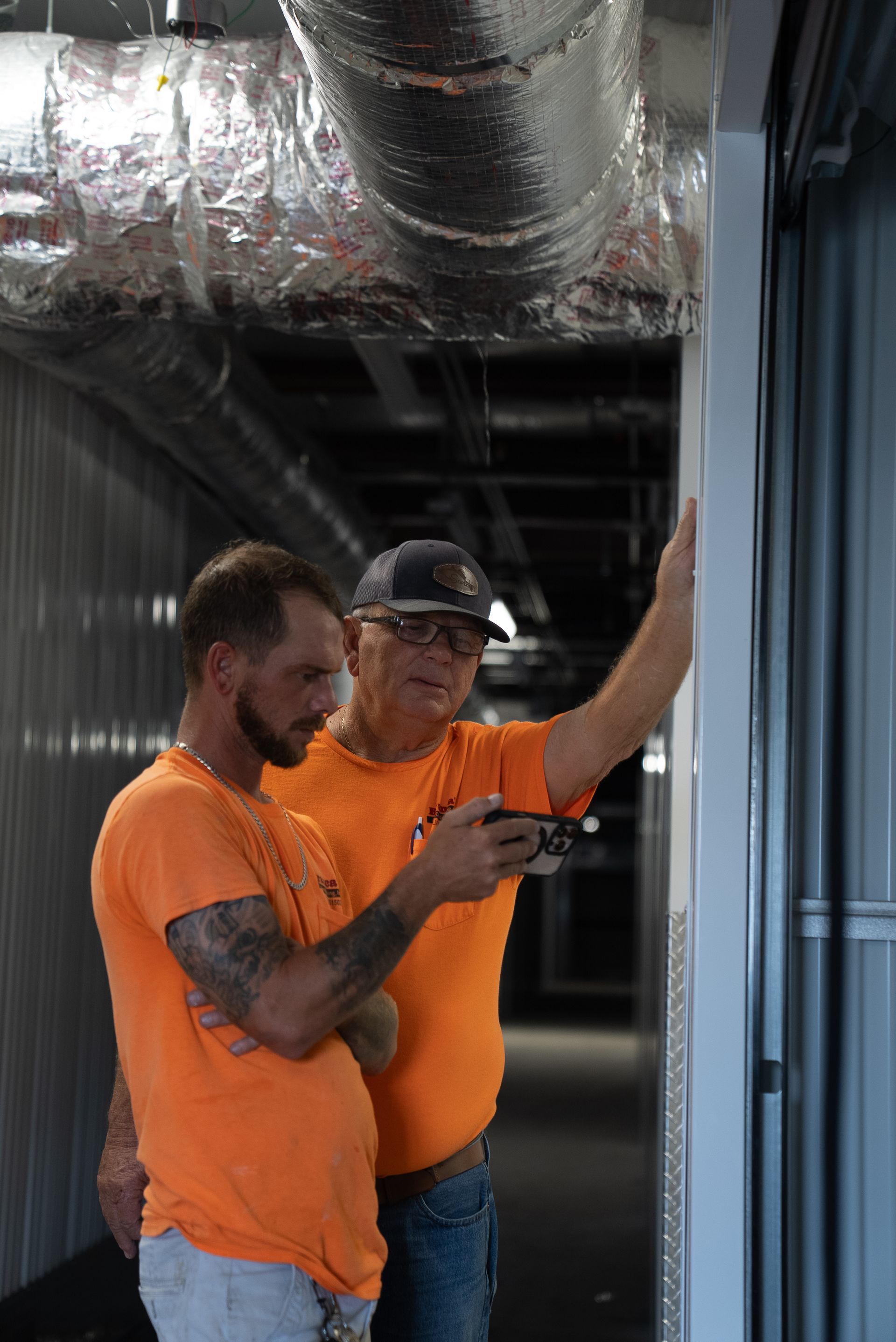 Two workers in orange shirts inspect metal framing in an unfinished building interior.