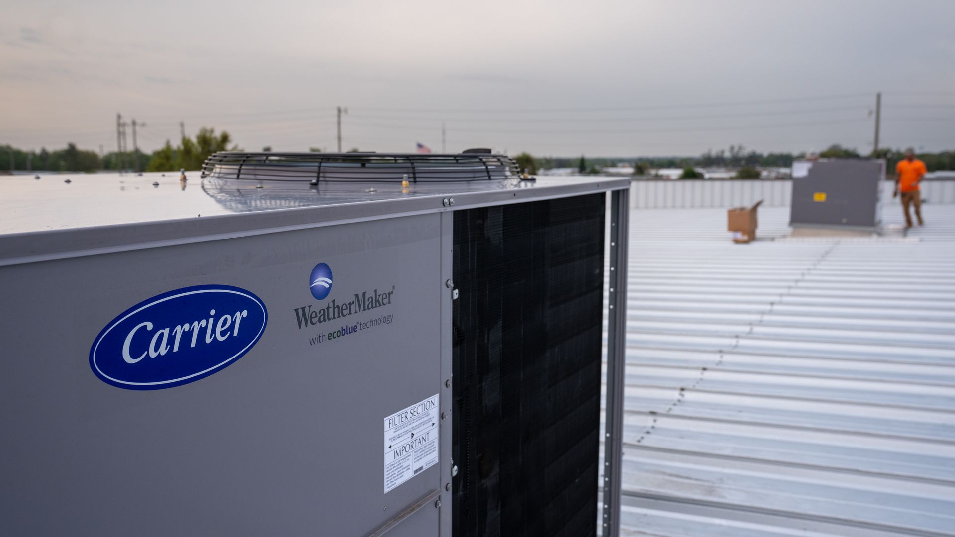 Carrier HVAC unit on a white metal rooftop with a worker in orange nearby at dusk