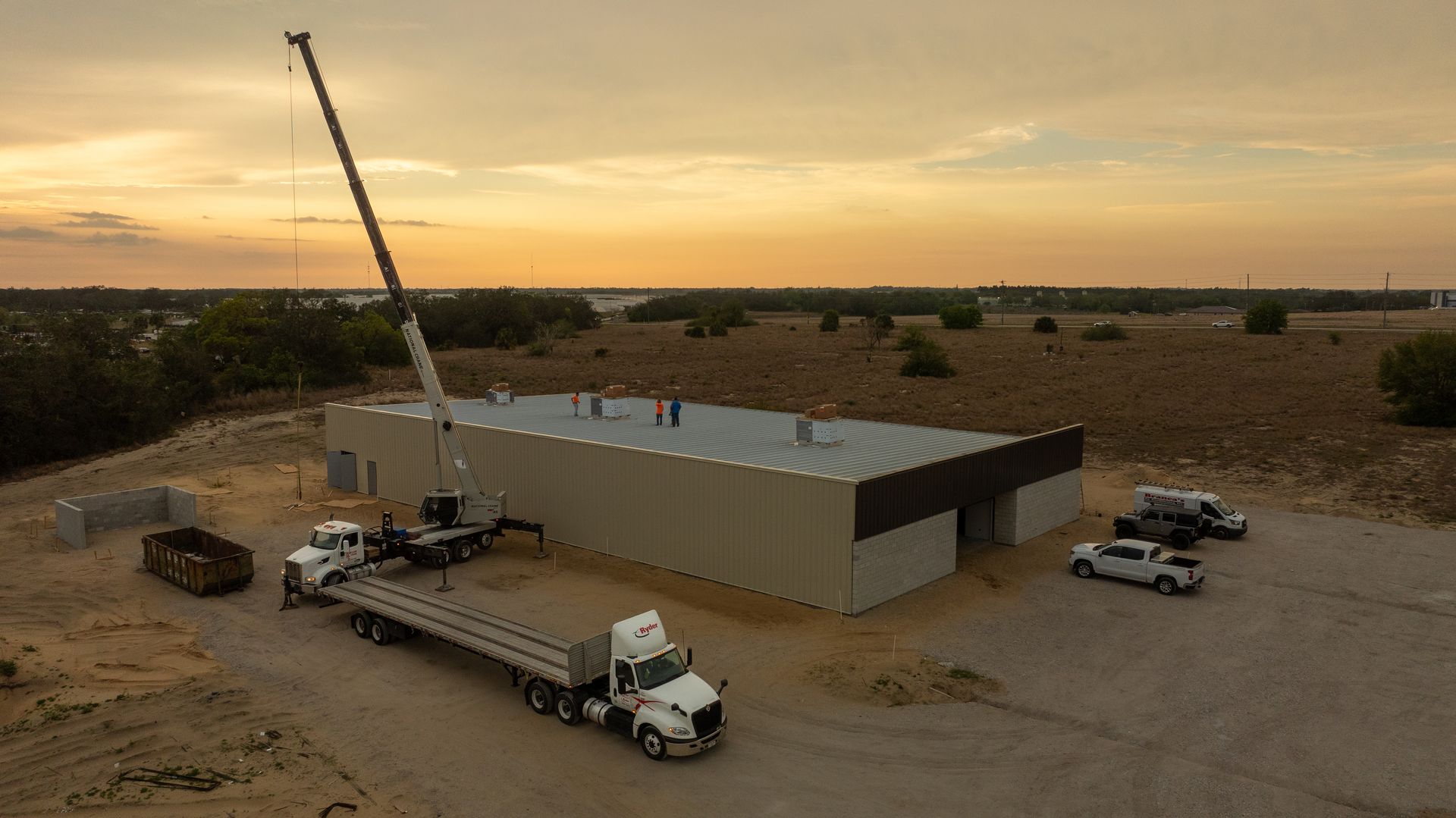 Crane lifting materials beside a long beige building in a rural dirt lot at sunset