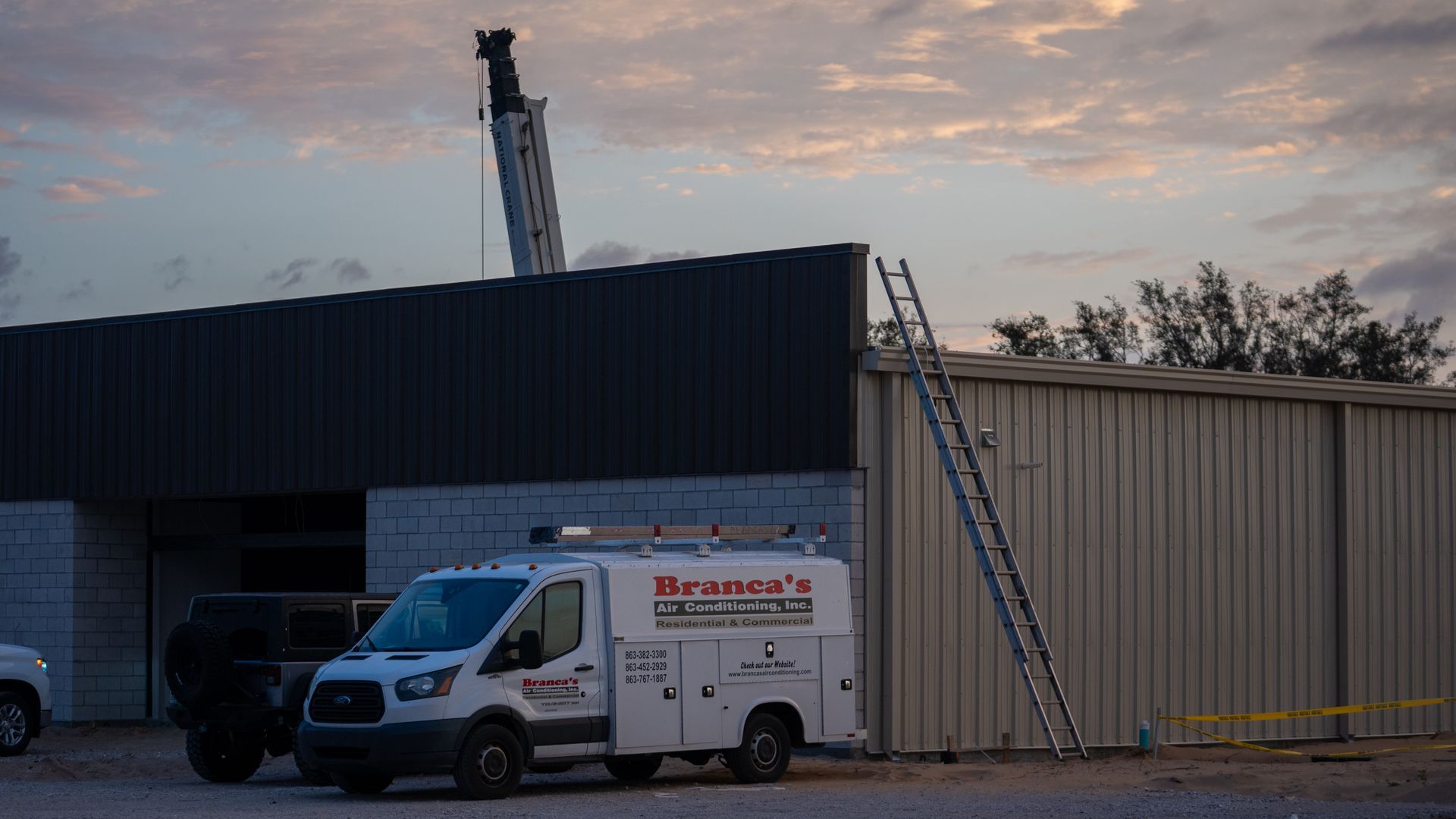Service van parked outside a metal building with a ladder against the wall at dusk.