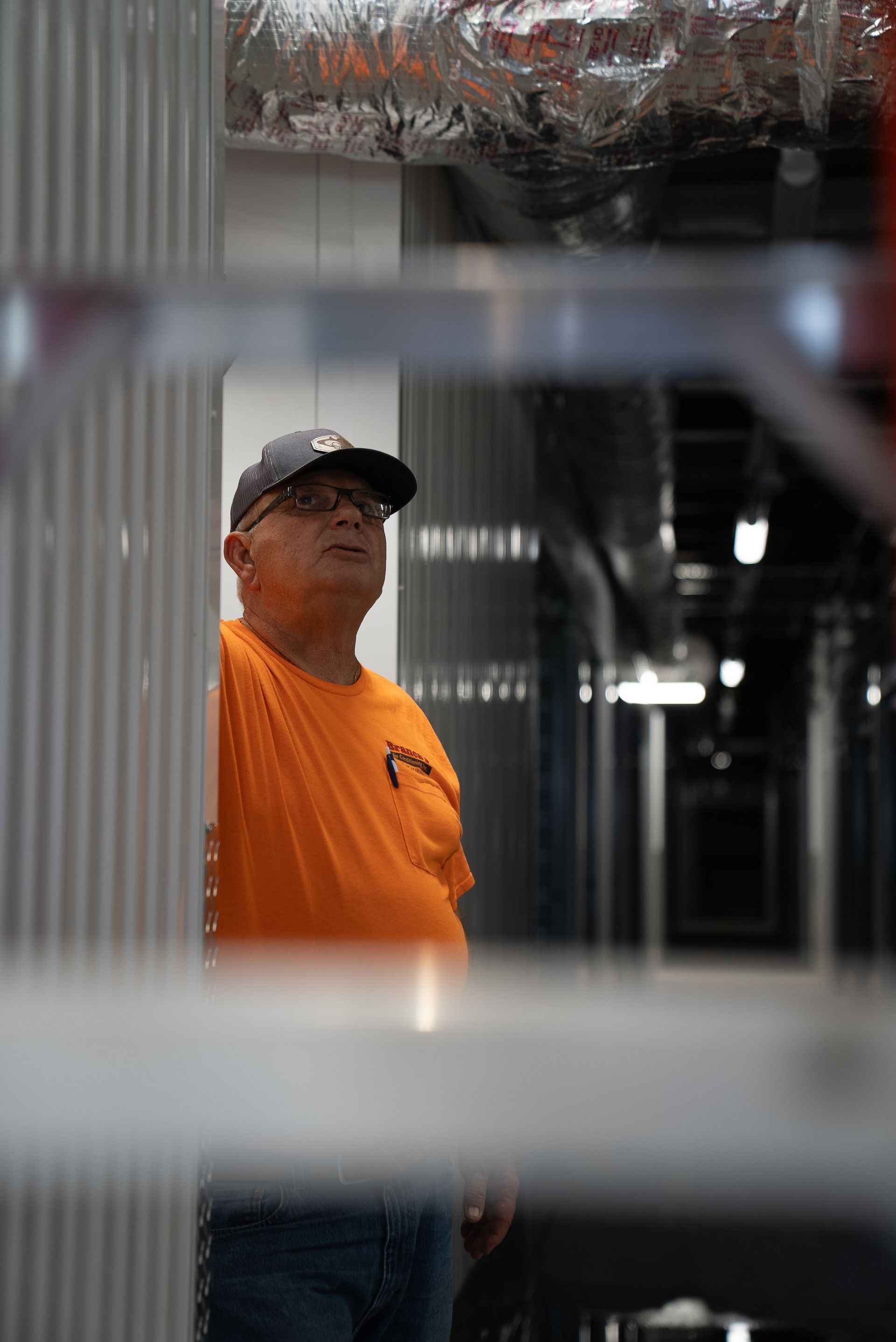 Worker in orange shirt and black cap standing in an industrial hallway, viewed through a metal frame