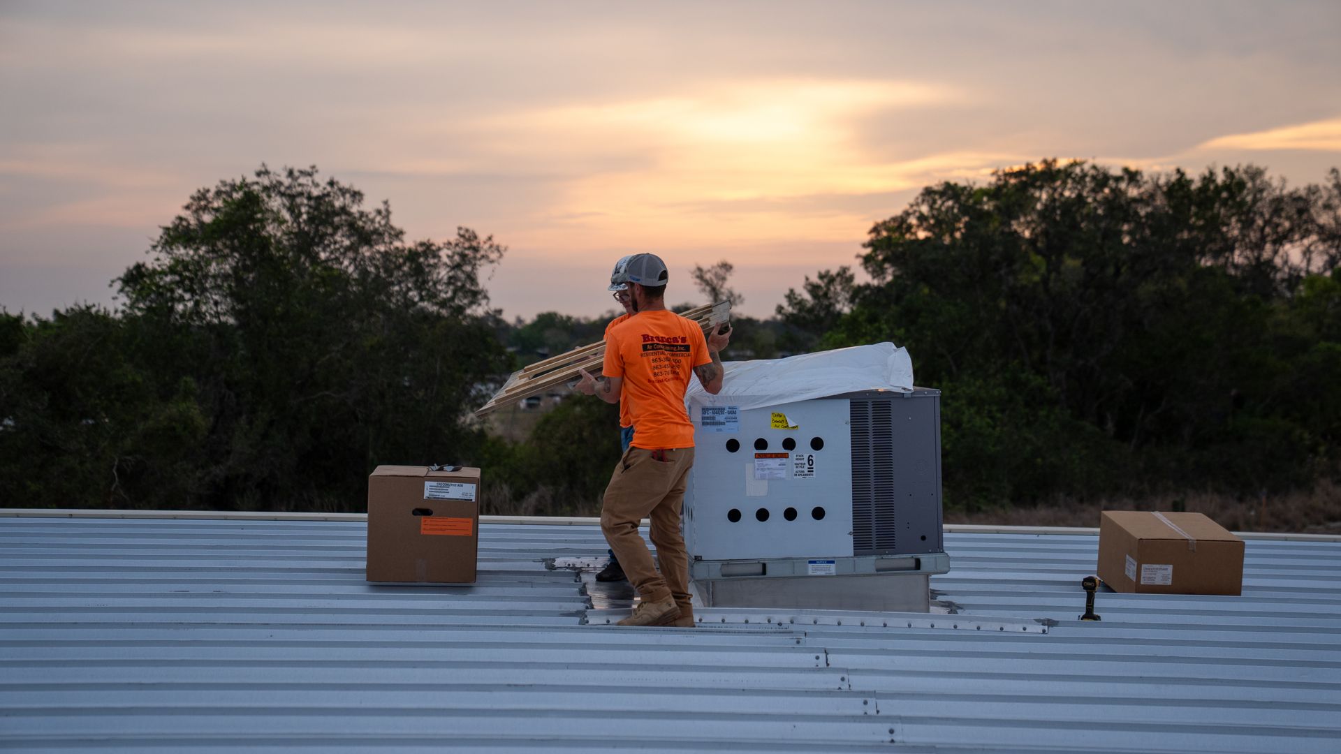 Worker in orange safety vest inspects rooftop equipment at sunset, with boxes nearby.
