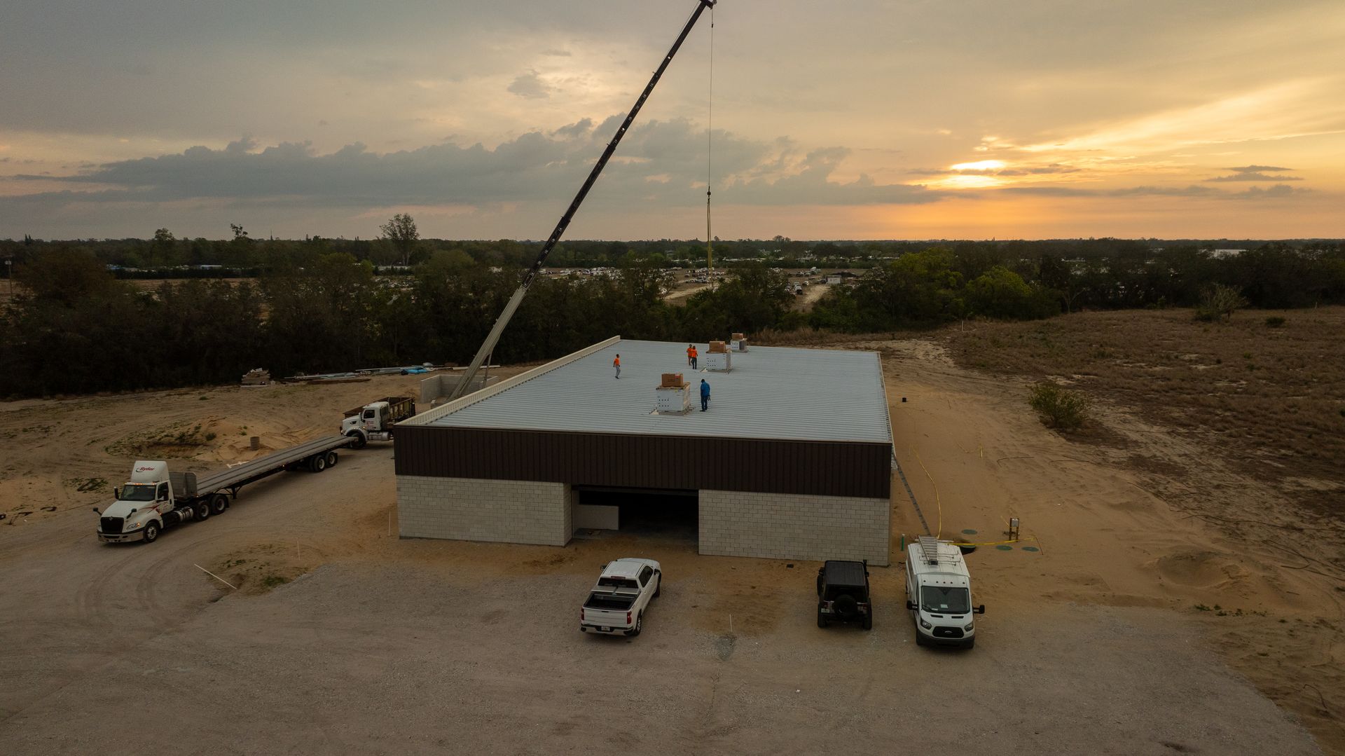 Construction site with a large metal building, crane, and vehicles at sunset