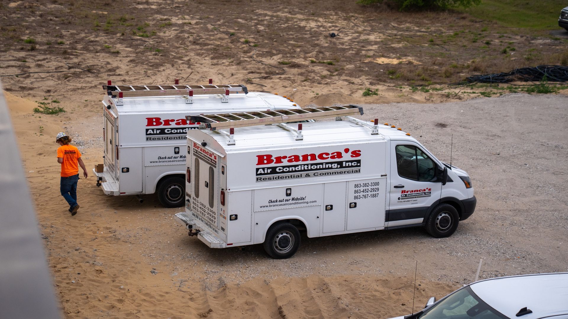 Two white service vans parked on dirt road beside a worker in orange safety gear